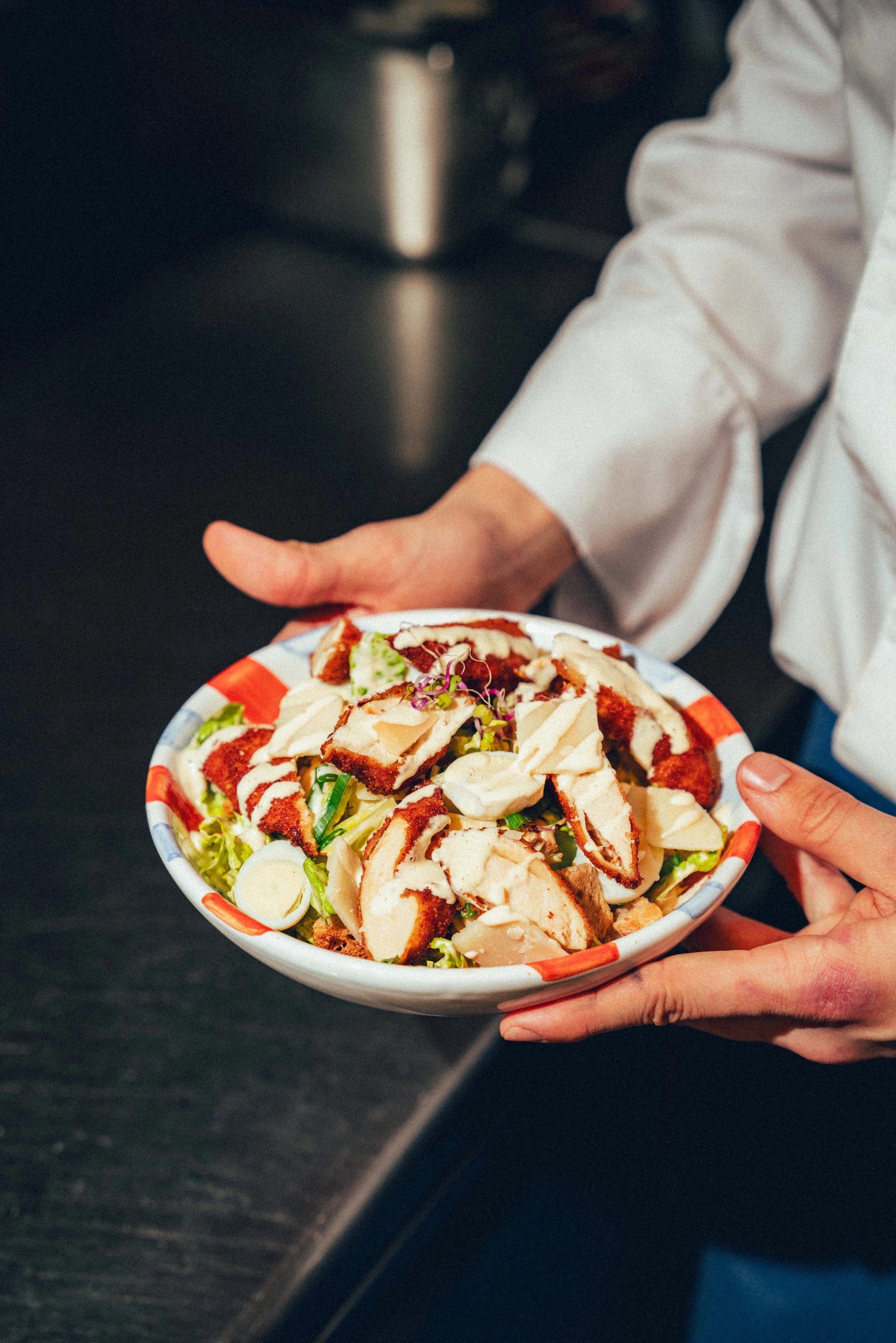 Une personne tient un bol de salade avec des morceaux de poulet grillé, des légumes et de la sauce, dans un environnement probablement de cuisine ou de restaurant.