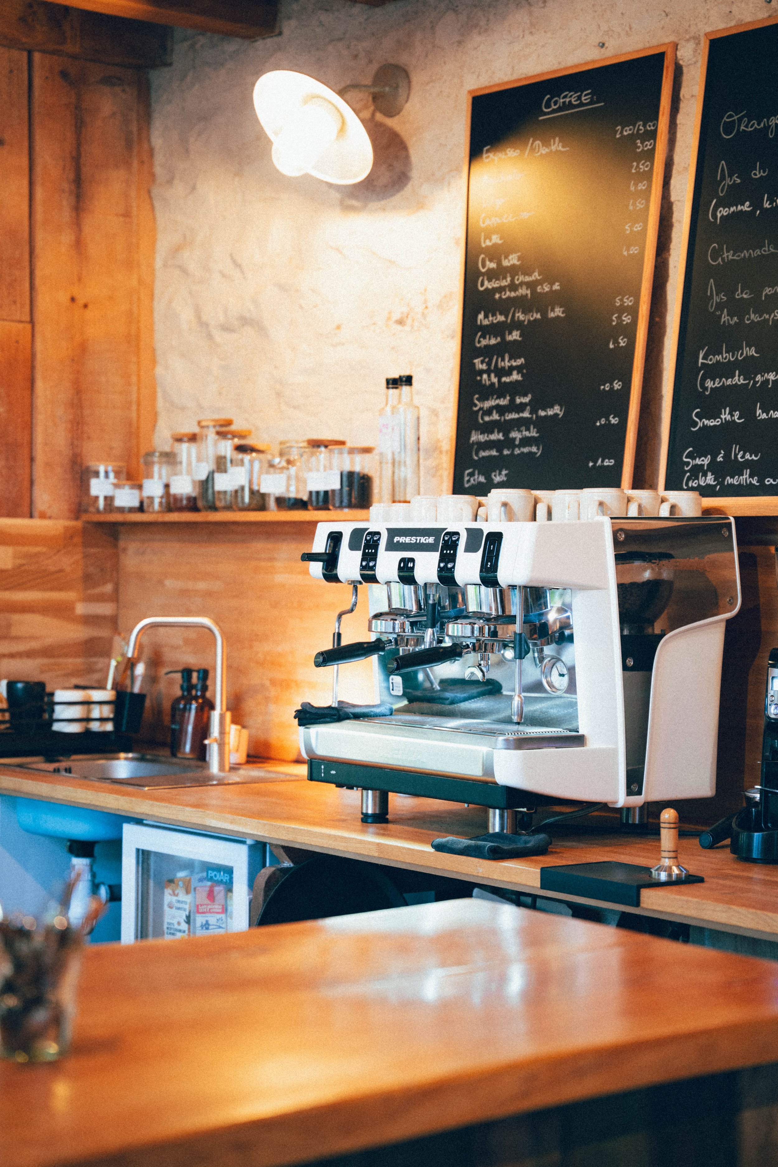 Une machine à café artisanale dans un café avec un tableau noir de menu en arrière-plan, étagères avec bocaux et espace de travail en bois.
