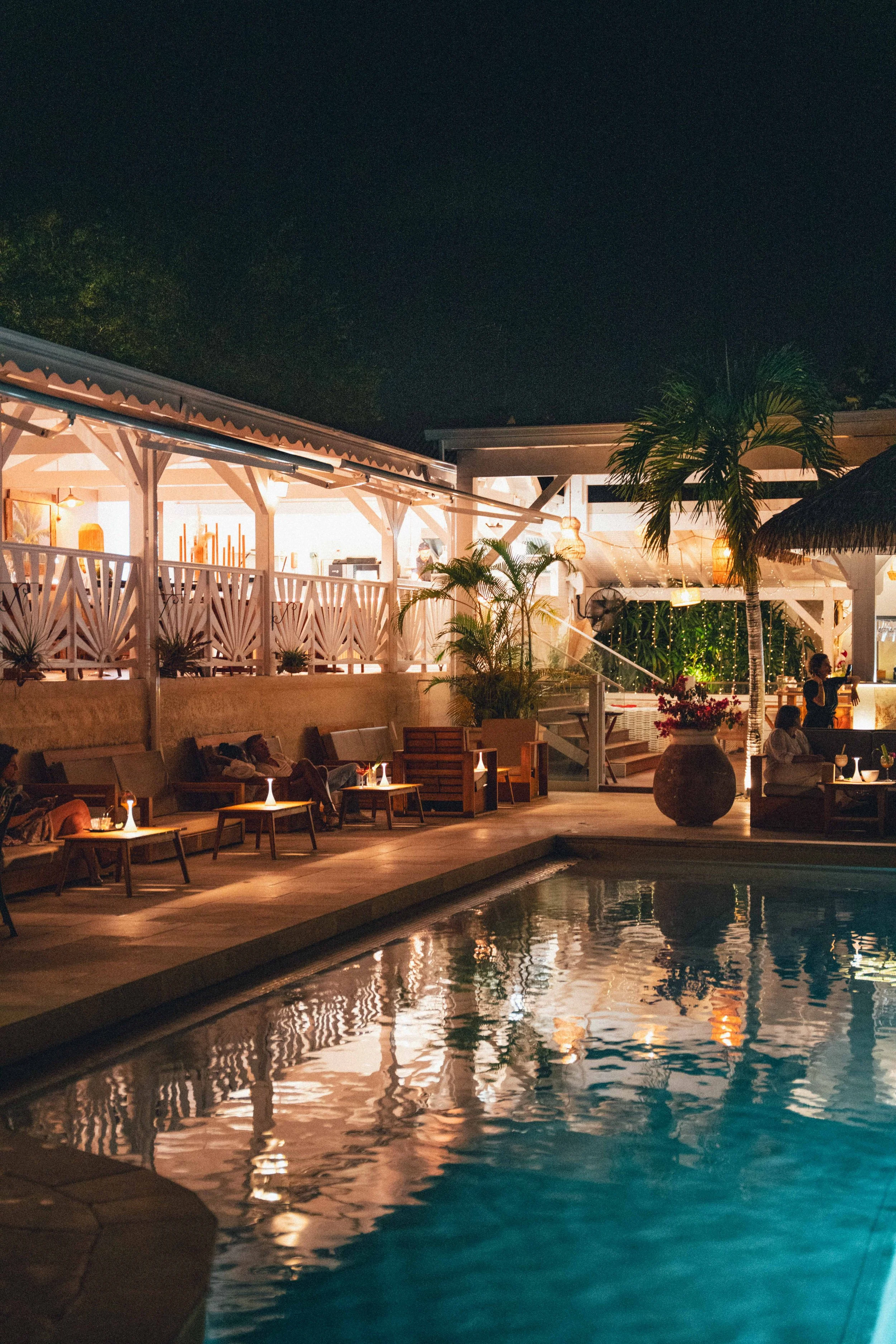 Piscine éclairée en soirée avec des chaises et tables en bois, des plantes tropicales, et une zone de restauration ou salon intérieur visible à l'arrière, ambiance chaleureuse et décontractée.