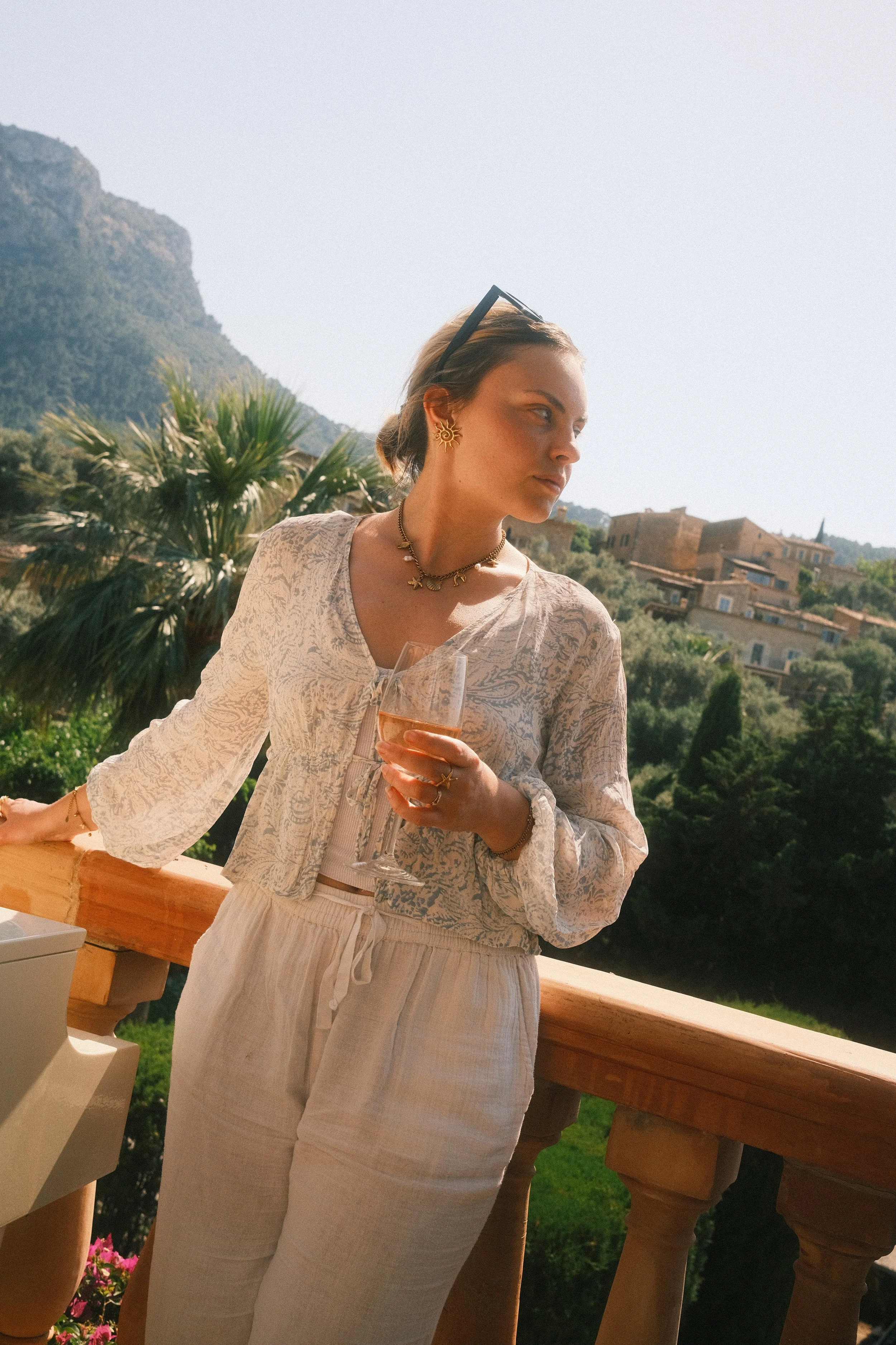 Femme blonde portant des bijoux et un vêtement beige, tenant un verre de vin rosé, en terrasse avec vue sur des montagnes et des maisons en arrière-plan.