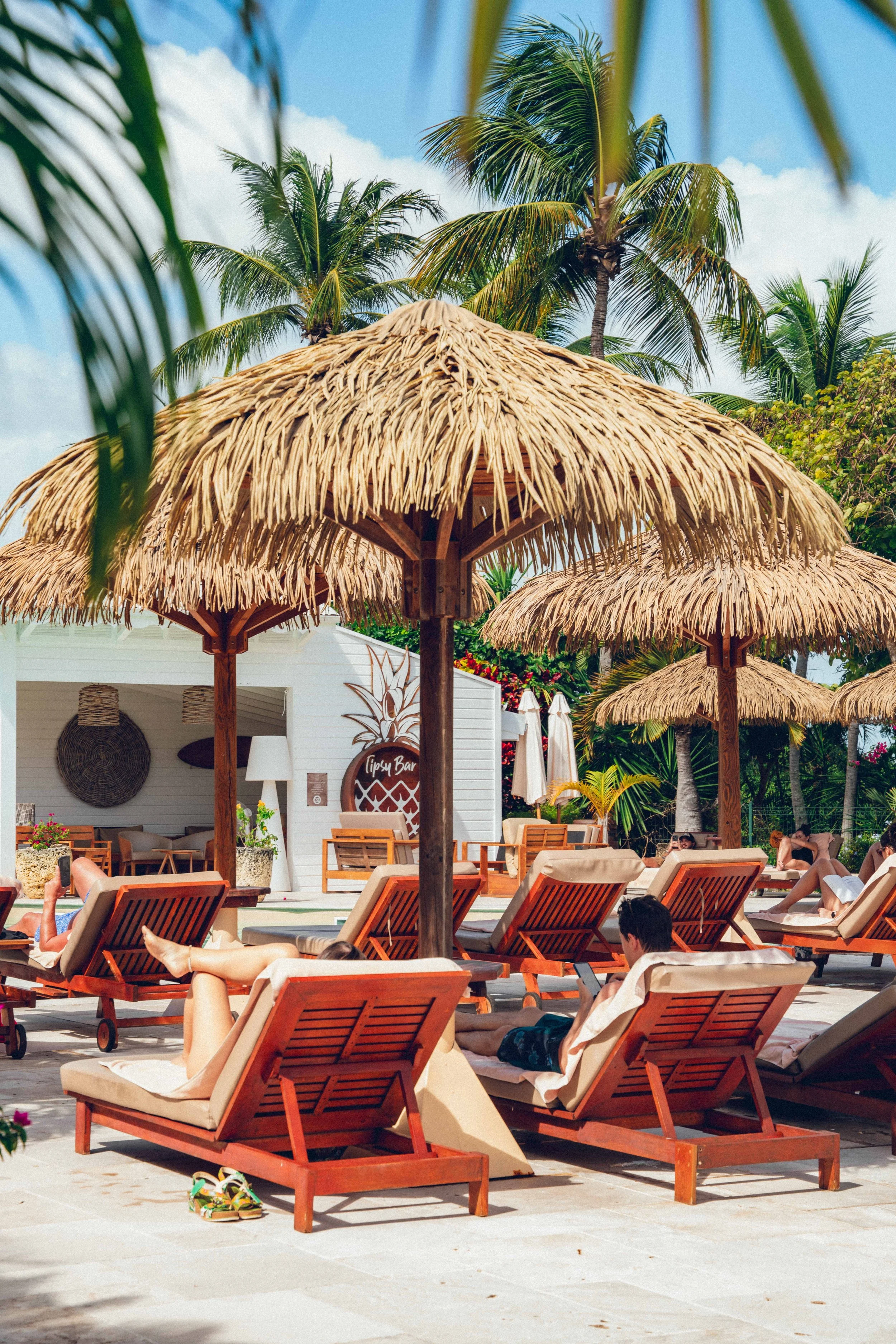 Transats en bois et parasols en chaume autour d'une piscine, avec des personnes allongées et se détendant, palmiers en arrière-plan, ambiance de vacances tropicales.