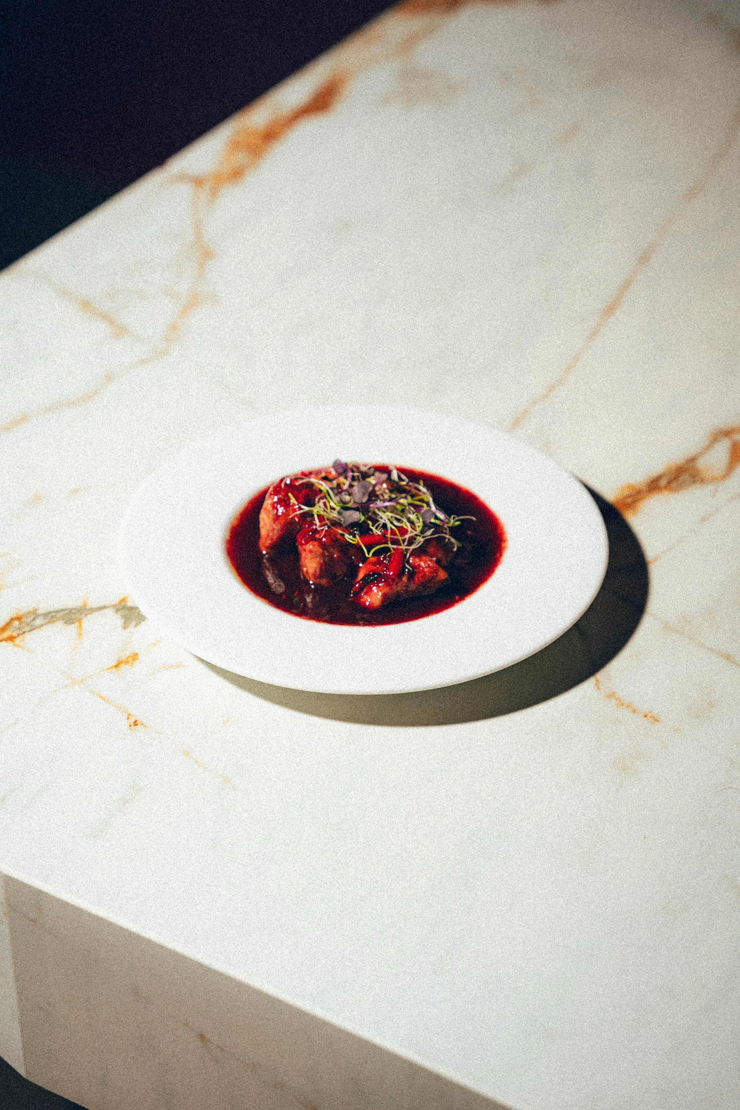 Plat de viande avec une sauce rouge, garniture de micro-pousses, sur une assiette blanche. Fond en marbre blanc avec veines dorées.