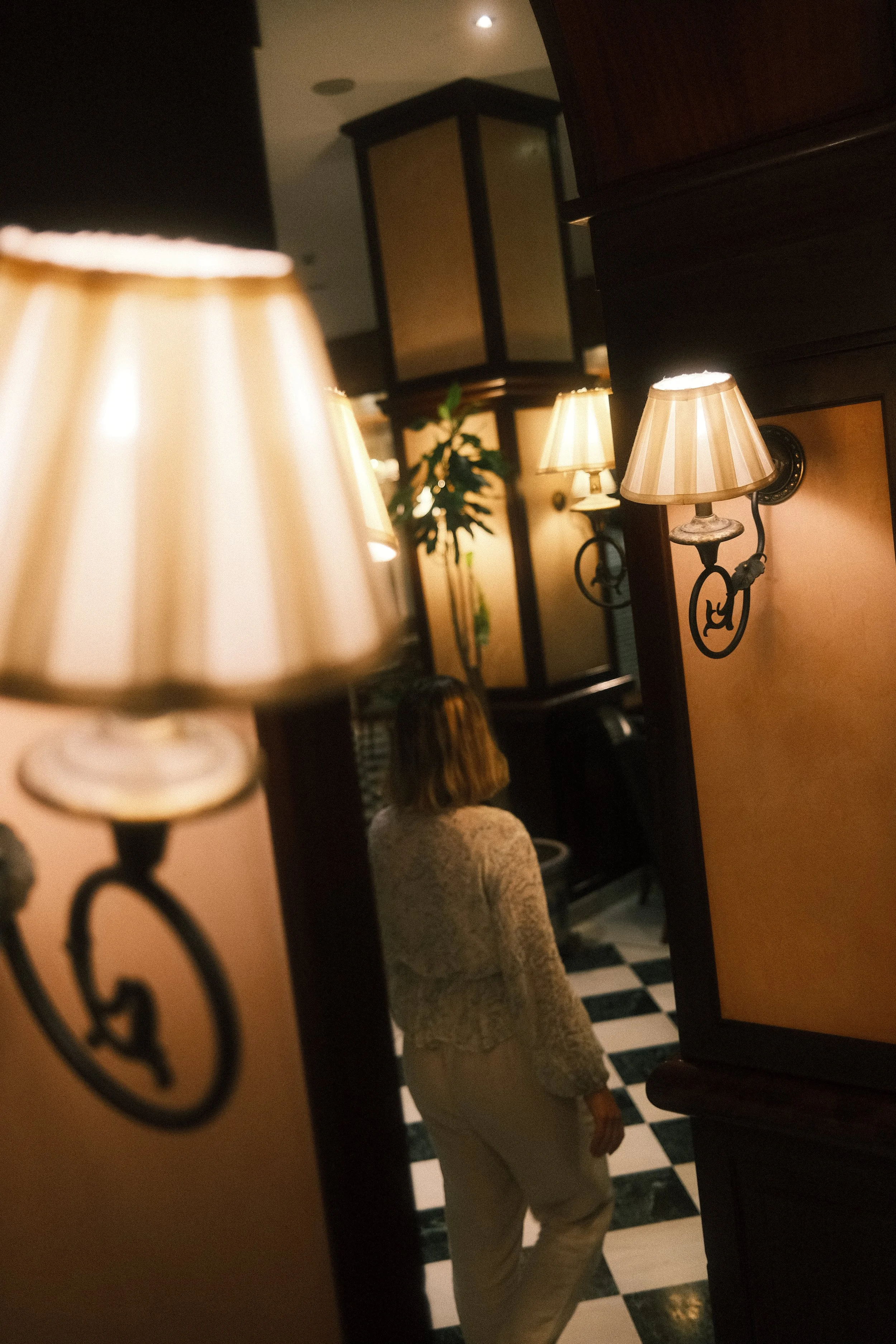 Une femme se tient dans un espace intérieur avec un sol à carreaux noirs et blancs, entourée de lampadaires à abat-jour beige, en regardant dans un miroir.