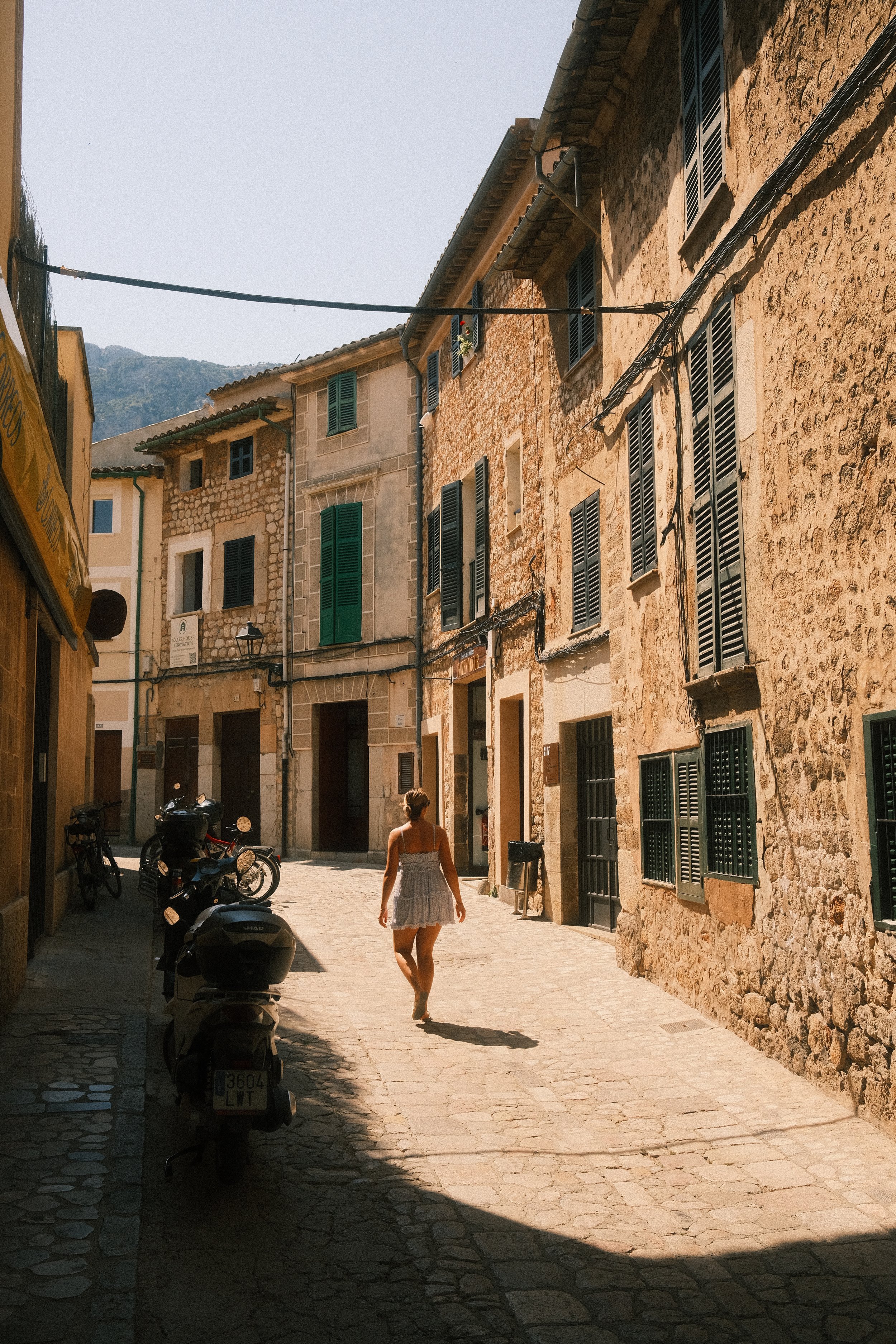 Une rue pavée bordée de bâtiments en pierre avec plusieurs fenêtres à volets verts et des motos garées sur le côté. Une femme en robe légère marche au centre, avec un arrière-plan de montagnes et un ciel clair.