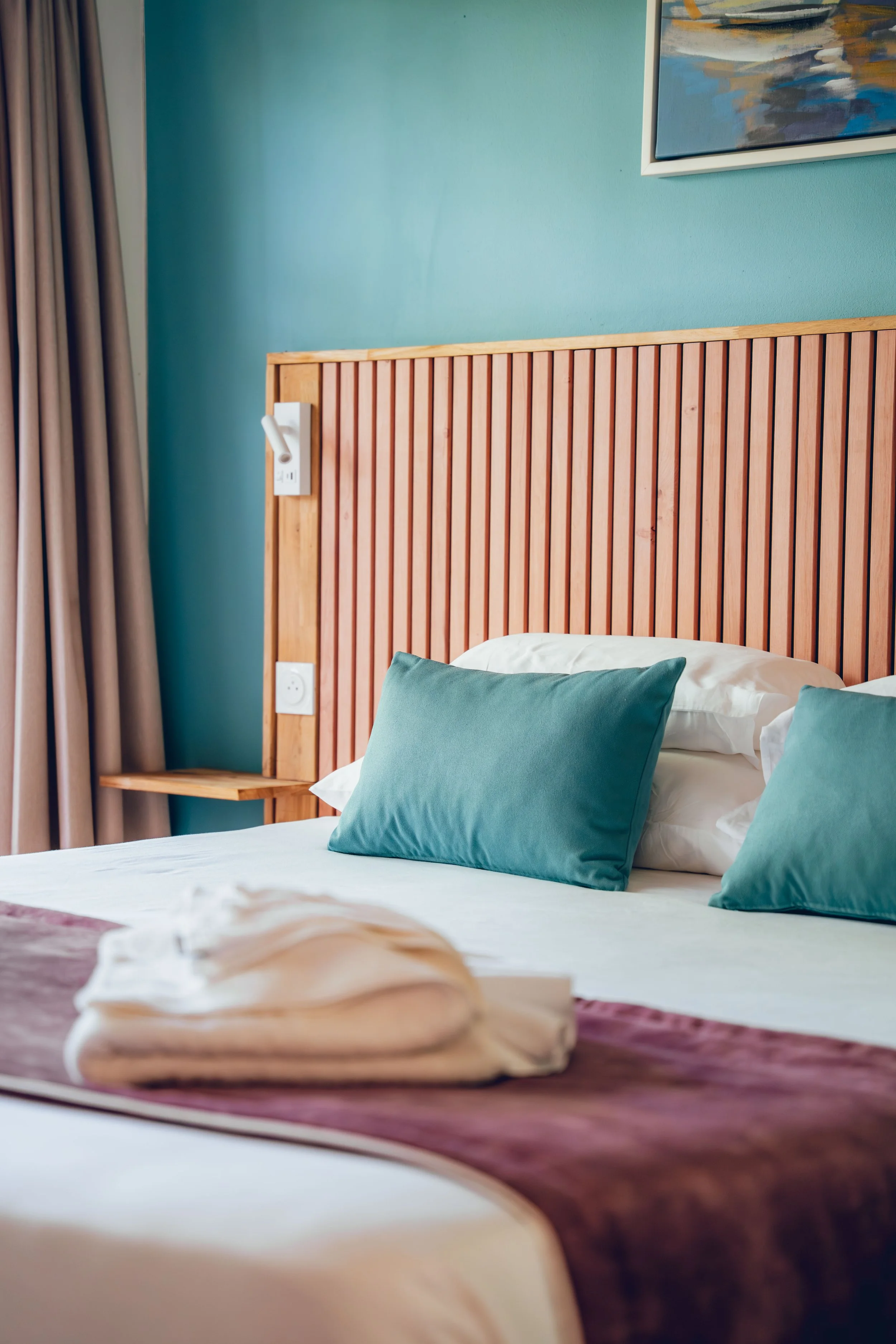 Photographie hôtelière d'un lit d'une chambre hôtelière avec des oreillers bleus, tête de lit en bois et rideaux beiges. Mur bleu avec une œuvre d'art abstraite.