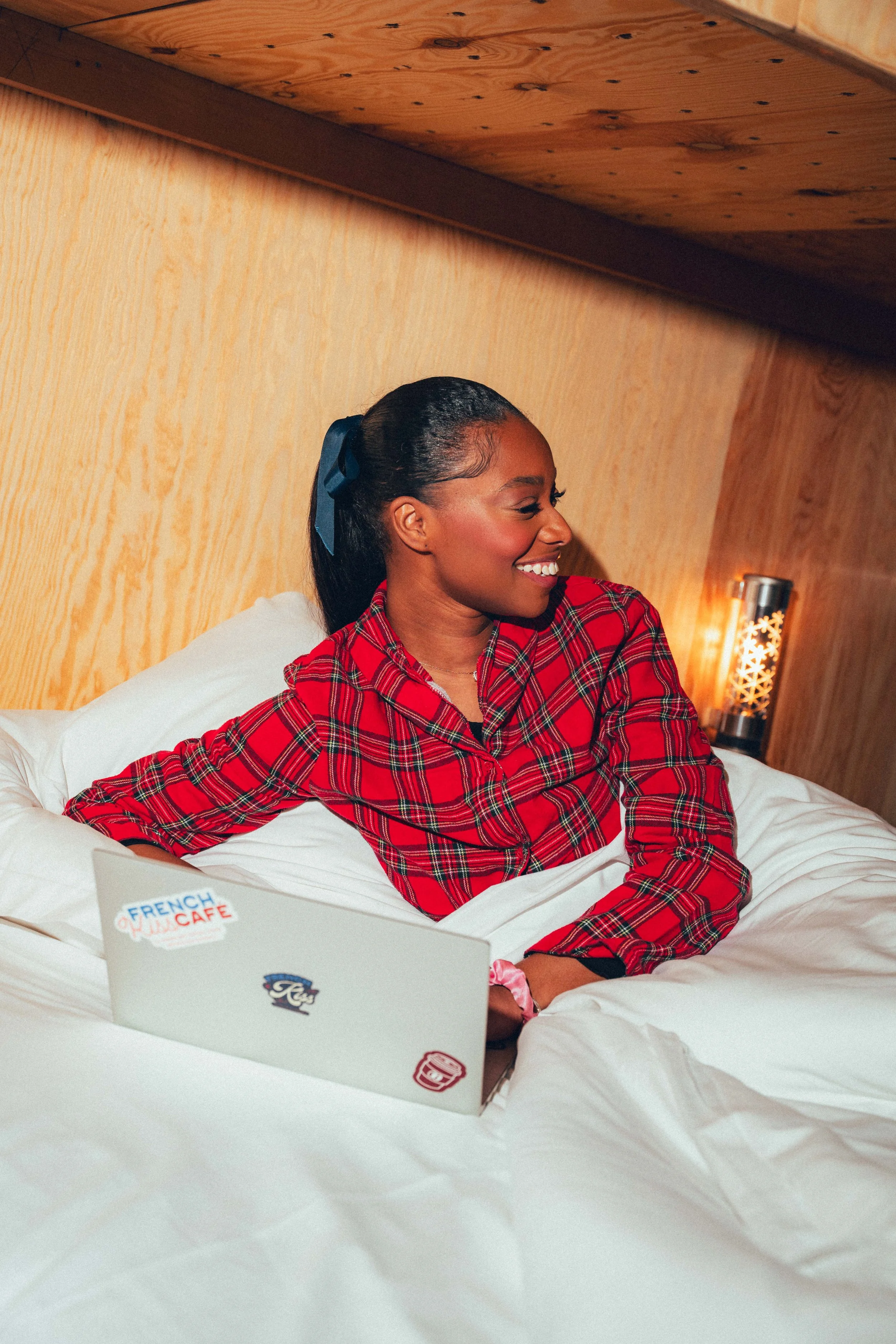 Une femme souriante à cheval sur un lit, portant une chemise à carreaux rouges et noirs, avec un ordinateur portable sur le lit. La pièce a un mur en bois et une lampe de chevet en arrière-plan.
