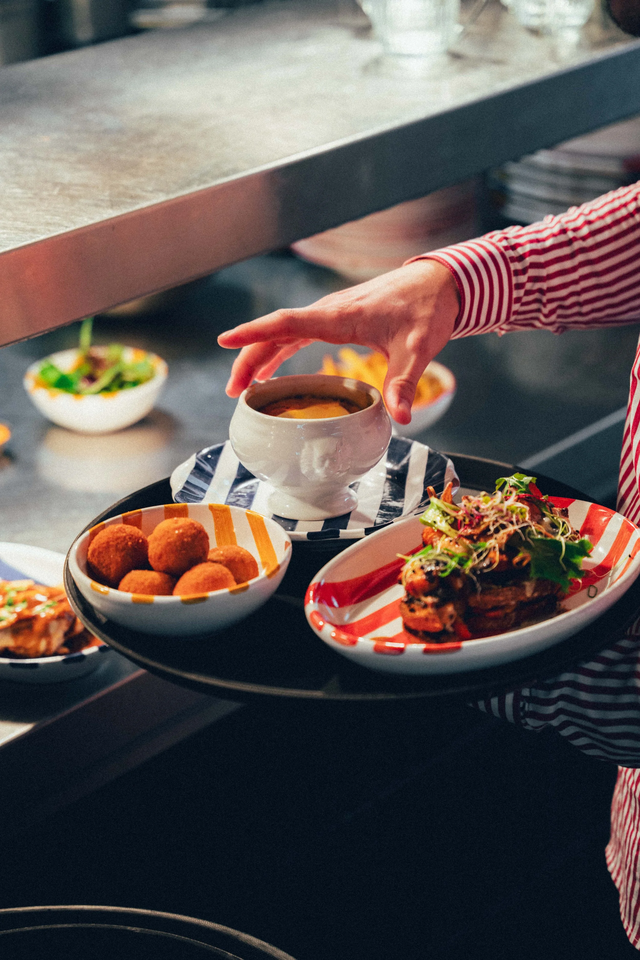 Une main place une tasse dans un environnement de cuisine avec un plateau de nourriture variée, comprenant des salades, des croquettes, et un plat principal, dans un cadre moderne.