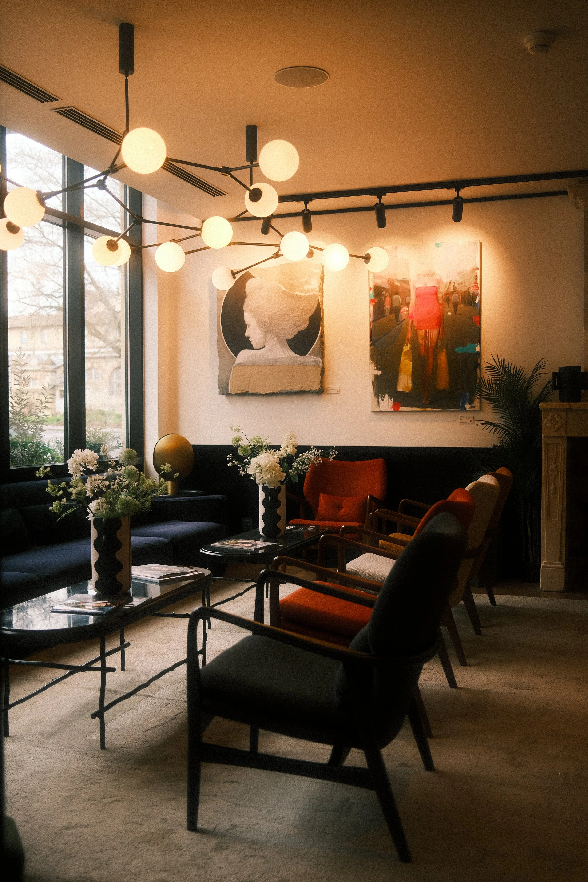Salon confortable avec des fauteuils, des tables basses, des vases contenant des fleurs, des œuvres d'art modernes accrochées aux murs, et un éclairage suspendu chaud, dans un intérieur lumineux grâce à de grandes fenêtres.