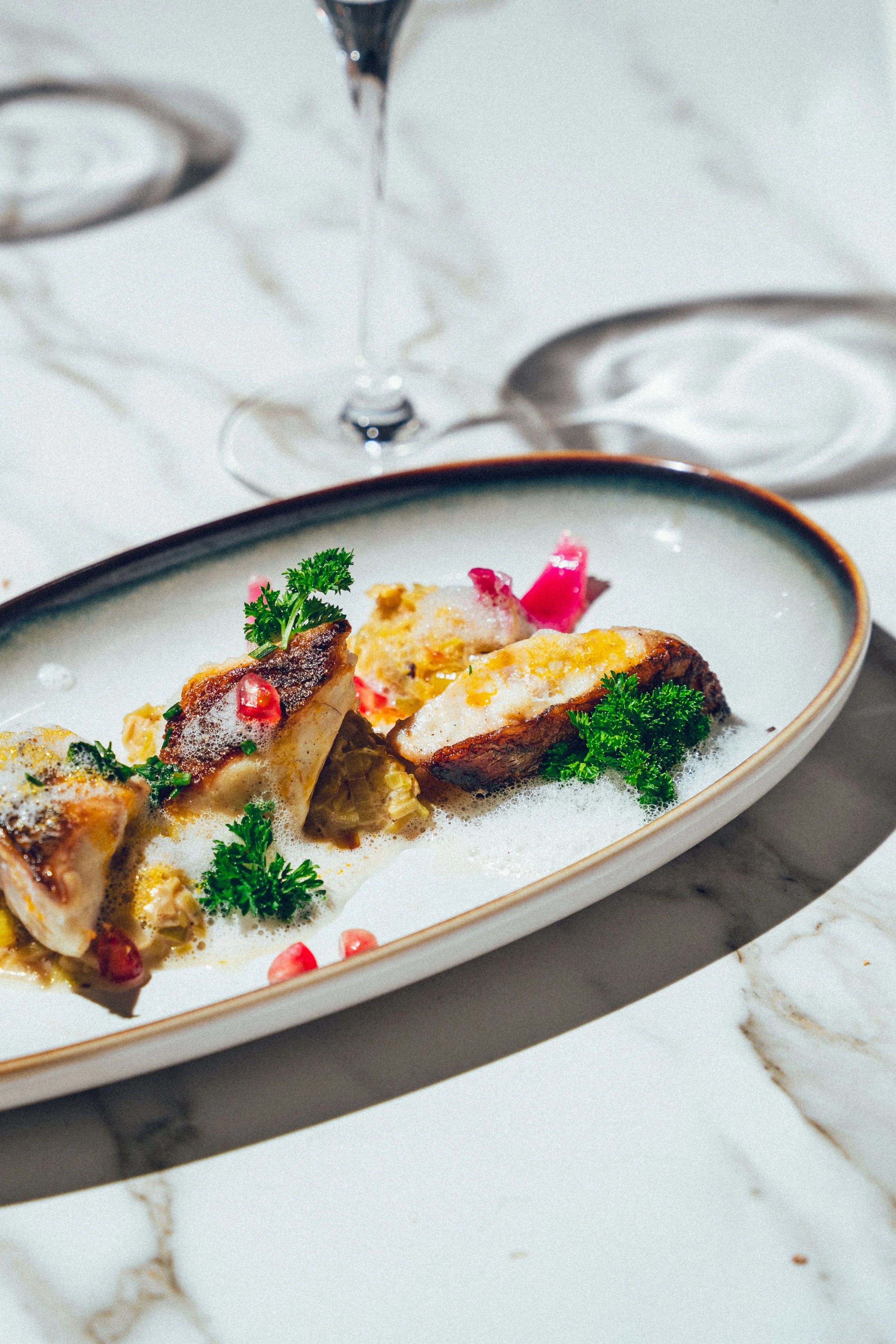 Photographie culinaire d'un plat gastronomique de poisson avec garnitures et herbes, servi dans une assiette blanche, avec des couverts en argent en arrière-plan sur une nappe blanche, sur une table en marbre.