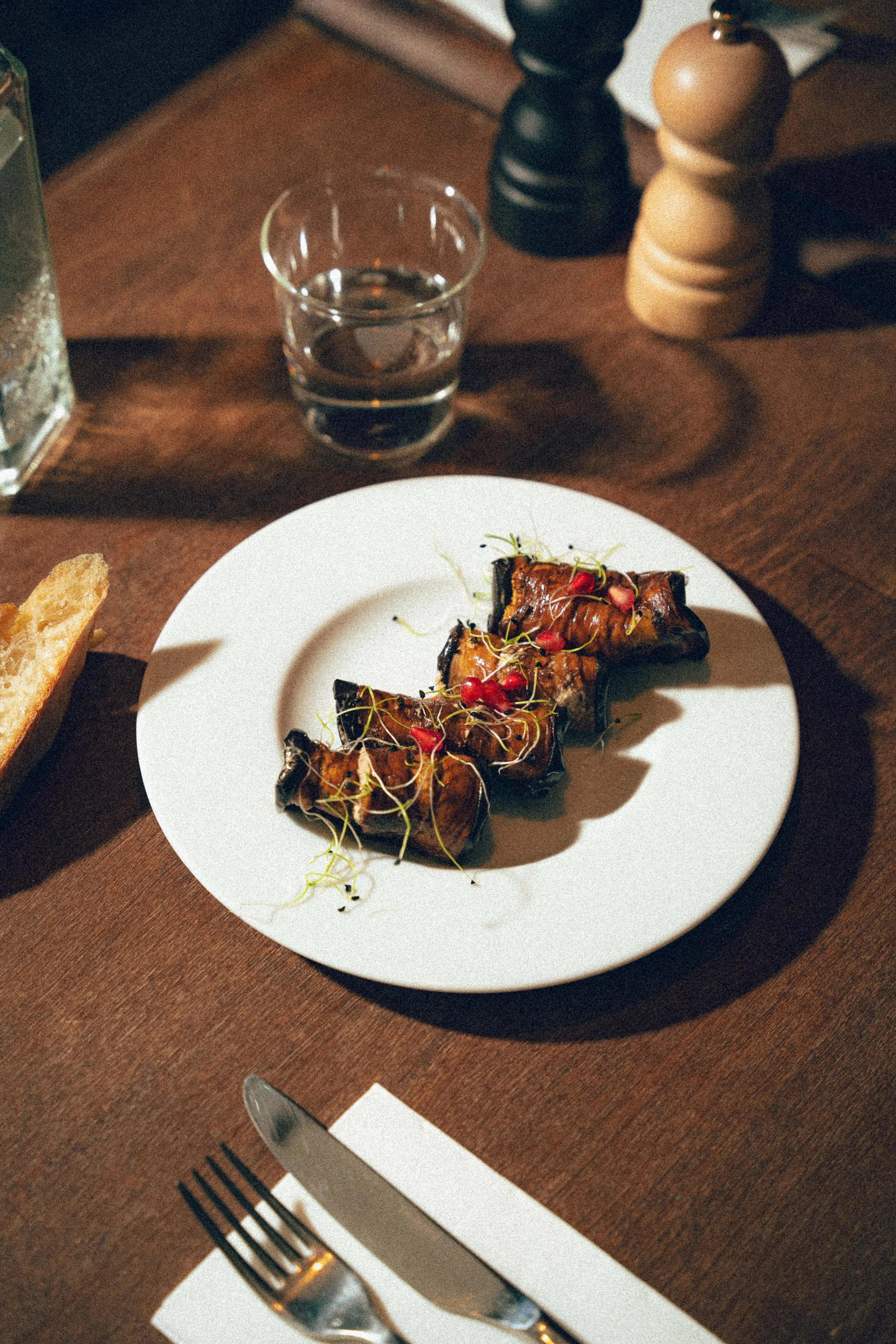 Une assiette avec des brochettes de légumes et de viande, garnies de micro-pousses et de petits fruits rouges, sur une table en bois. En arrière-plan, un verre d'eau, des salières en bois et un couteau et une fourchette posés sur une nappe blanche.