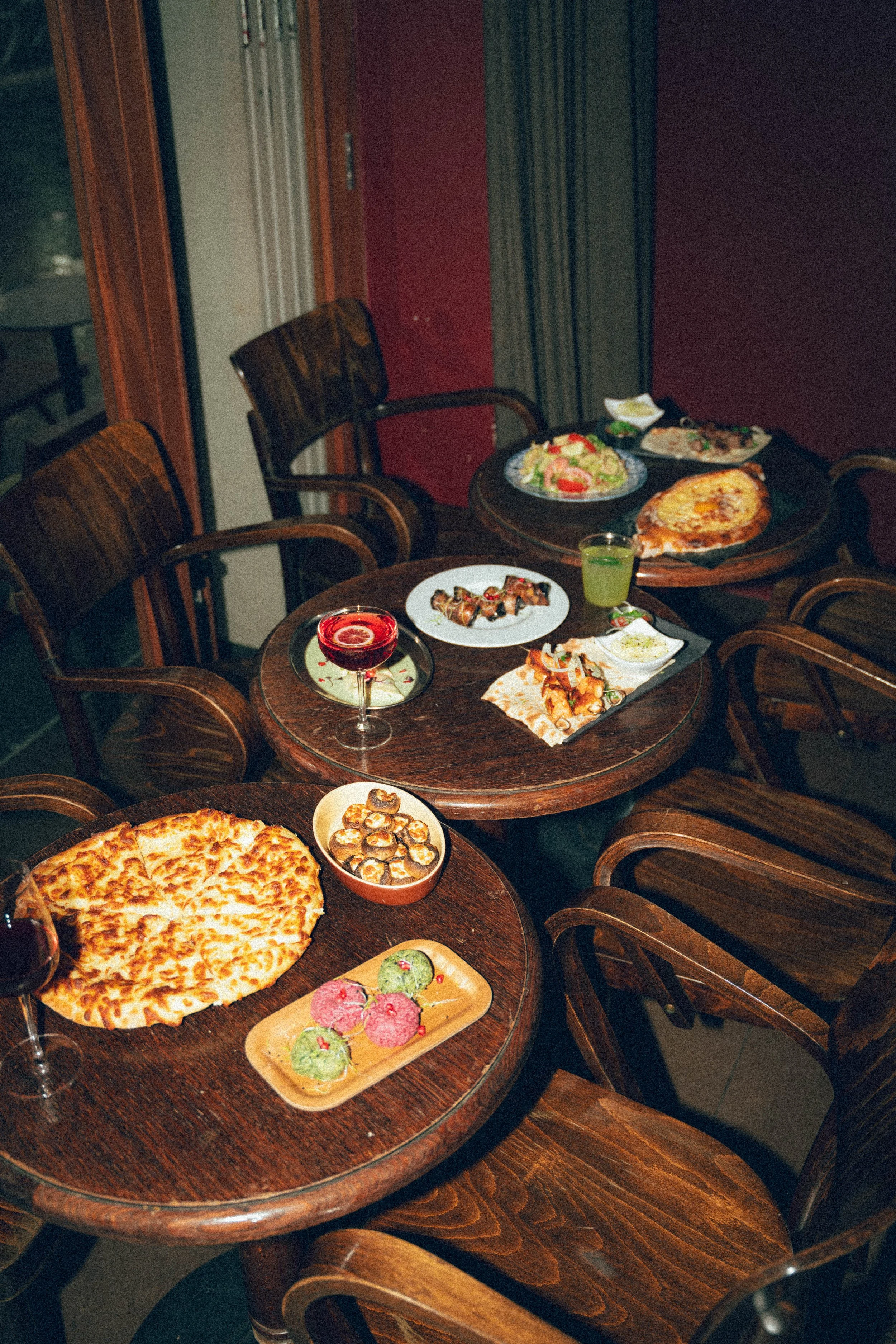 Table avec plusieurs plats de nourriture dont une pizza, une salade, un verre de vin rouge, un cocktail vert, des sushis, des chips et des dips, dans un restaurant.