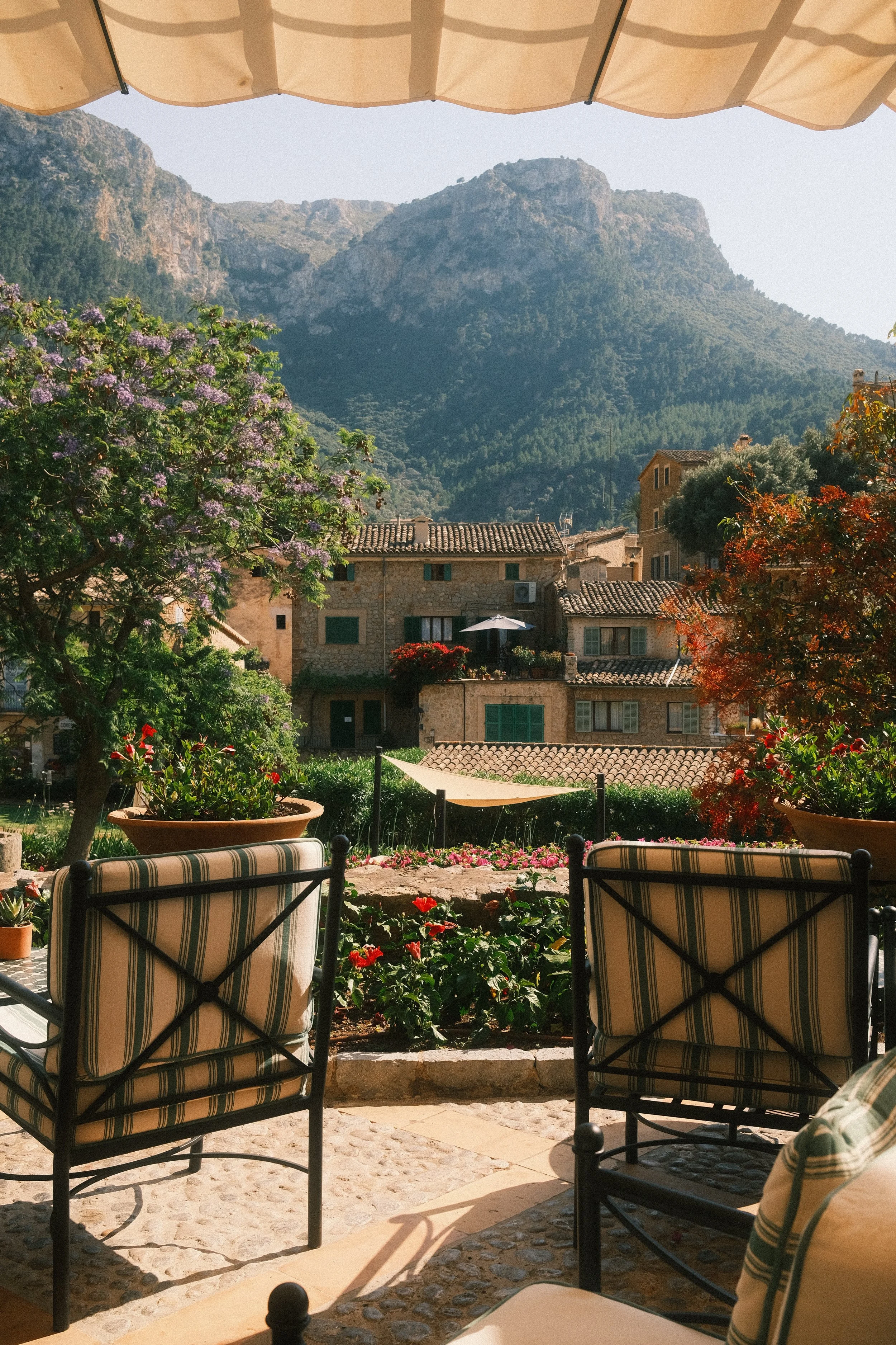 Une vue d'une terrasse avec des chaises et des plantes, donnant sur un village en pierre avec des montagnes en arrière-plan. Un parasol beige couvre la terrasse.