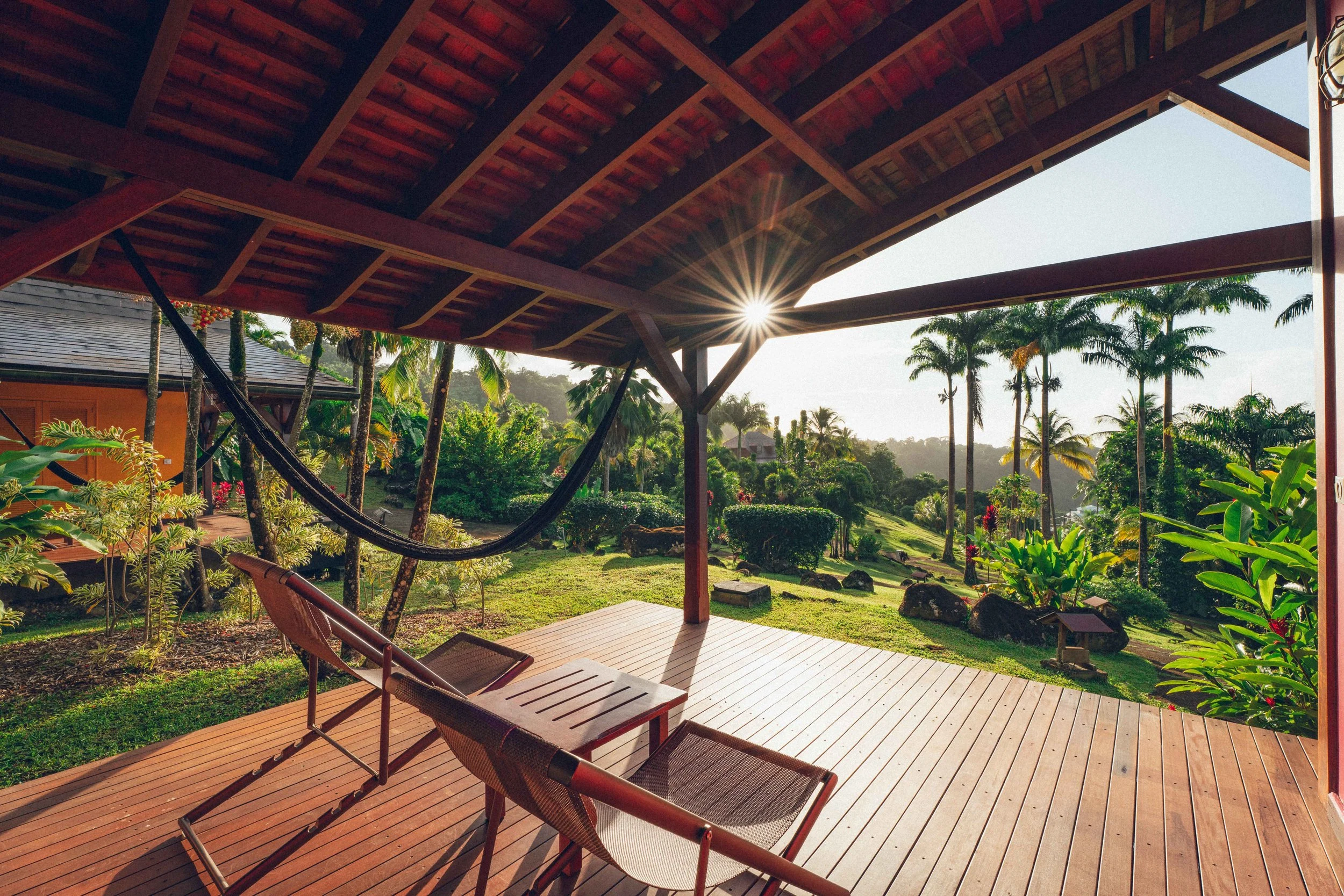 Une terrasse en bois avec deux chaises longues en regardant un jardin tropical avec des palmiers et d'autres plantes luxuriantes sous un ciel ensoleillé.