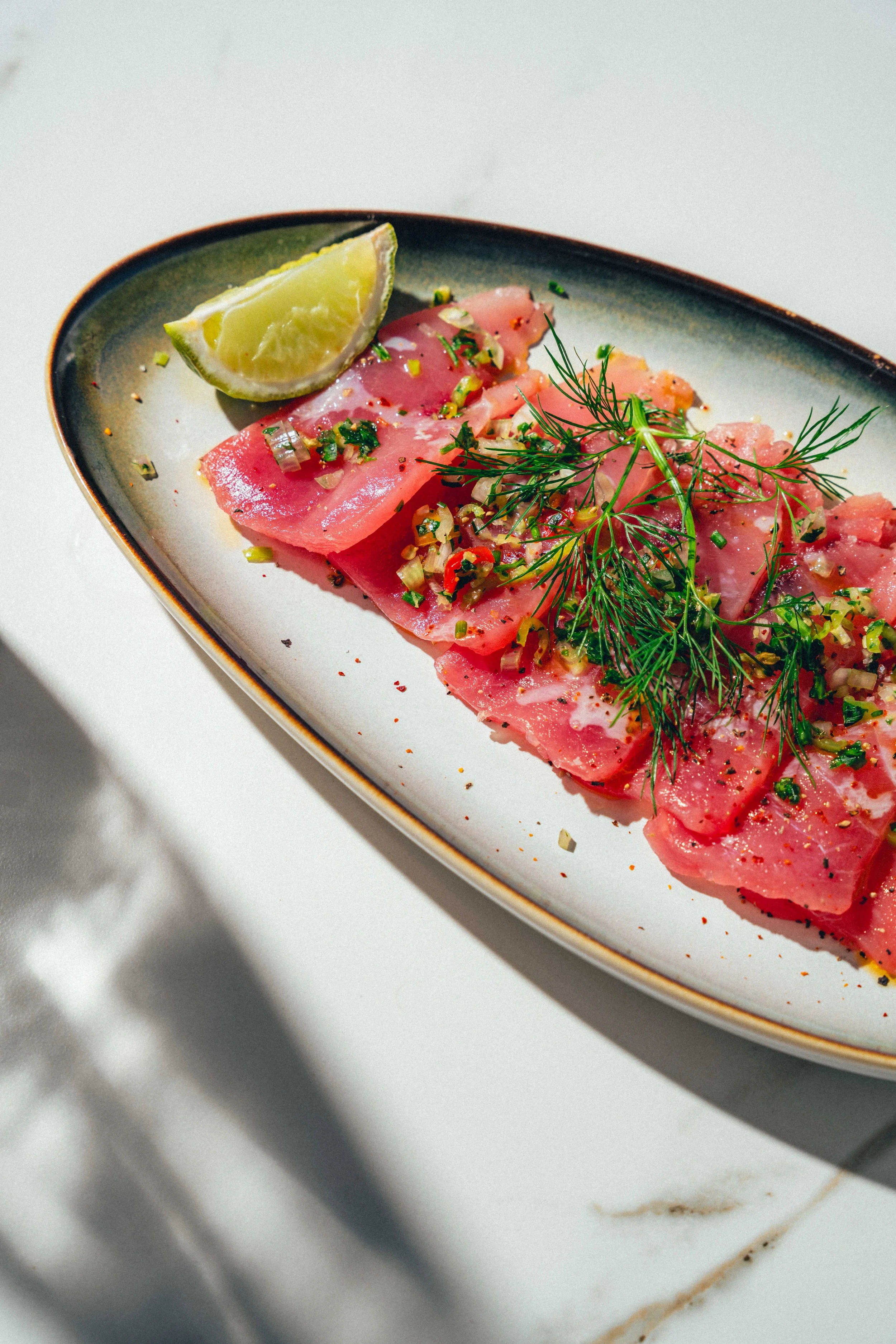 Photographie d'un plat gastronomique de carpaccio de poisson garnie de fines herbes, d'oignons hachés, de sel, de poivre, et accompagnée d'un quartier de citron vert.