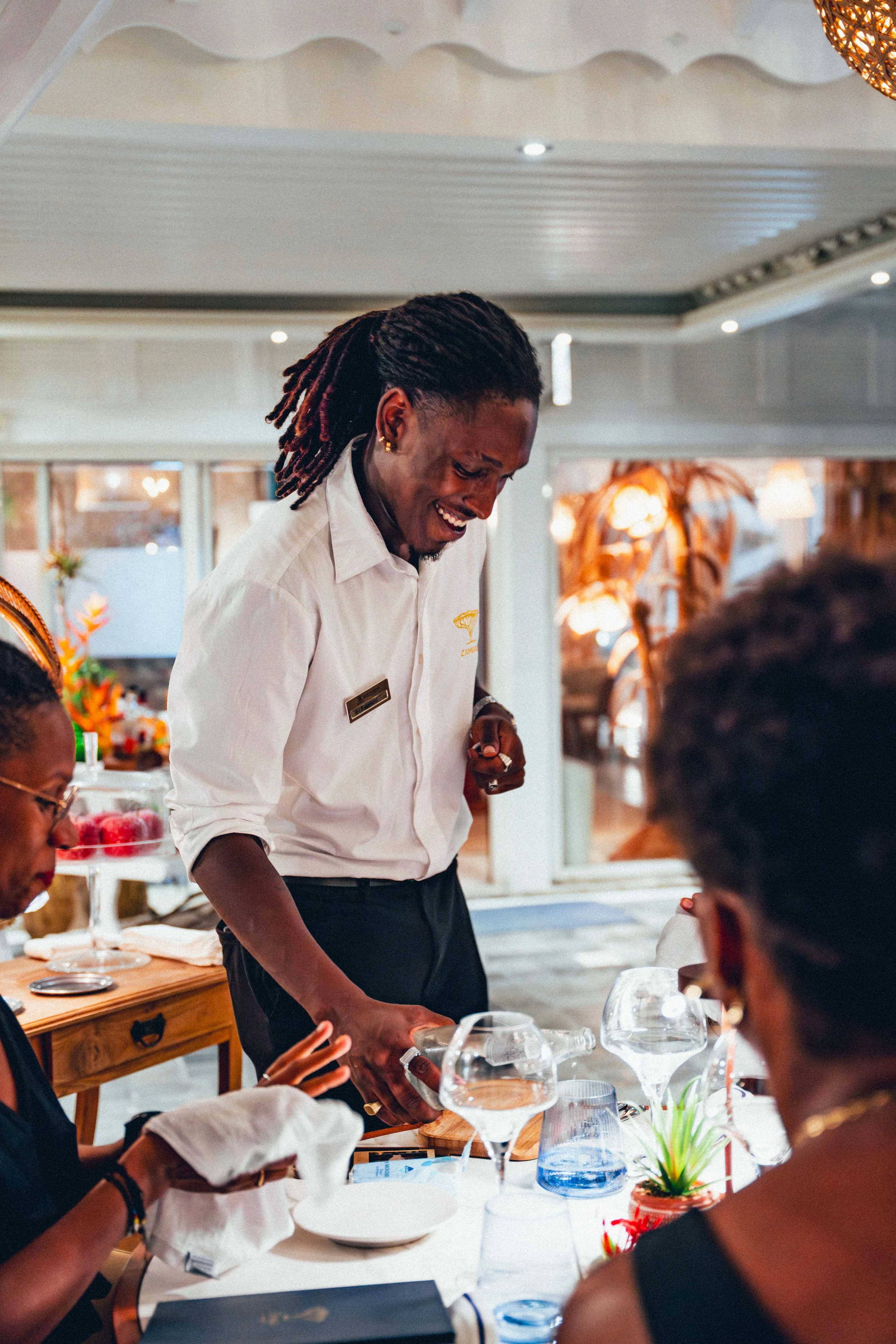 Photographie culinaire d'un serveur souriant en chemise blanche sert de la nourriture à une table de personnes lors d'un dîner en intérieur, décoré avec des éléments en bois et des éclairages chaleureux.