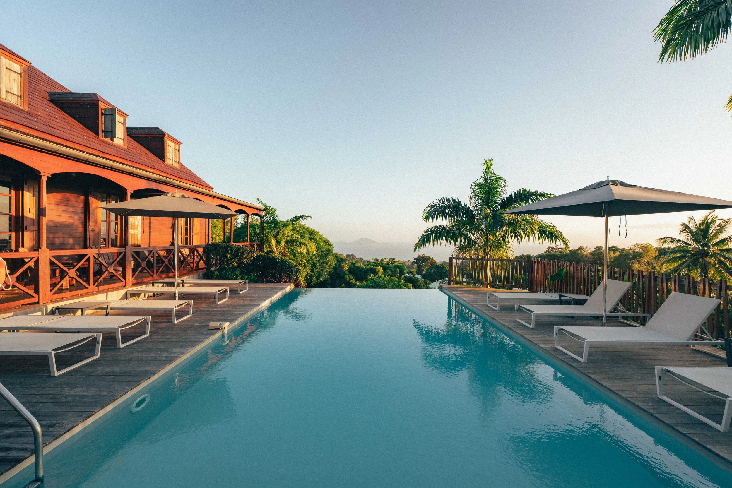 Piscine à débordement avec des chaises longues, parapluies et végétation tropicale, bâtiment en bois, vue sur la nature au coucher du soleil.