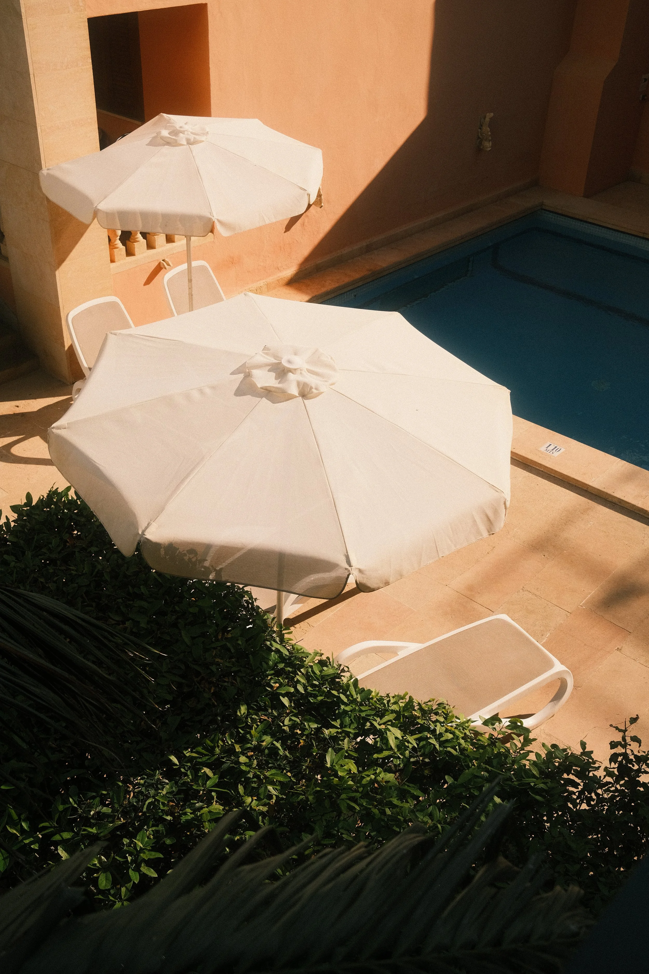 Deux parasols blancs ouverts à côté d'une piscine. La scène est en plein jour avec des ombres portées sur le sol.