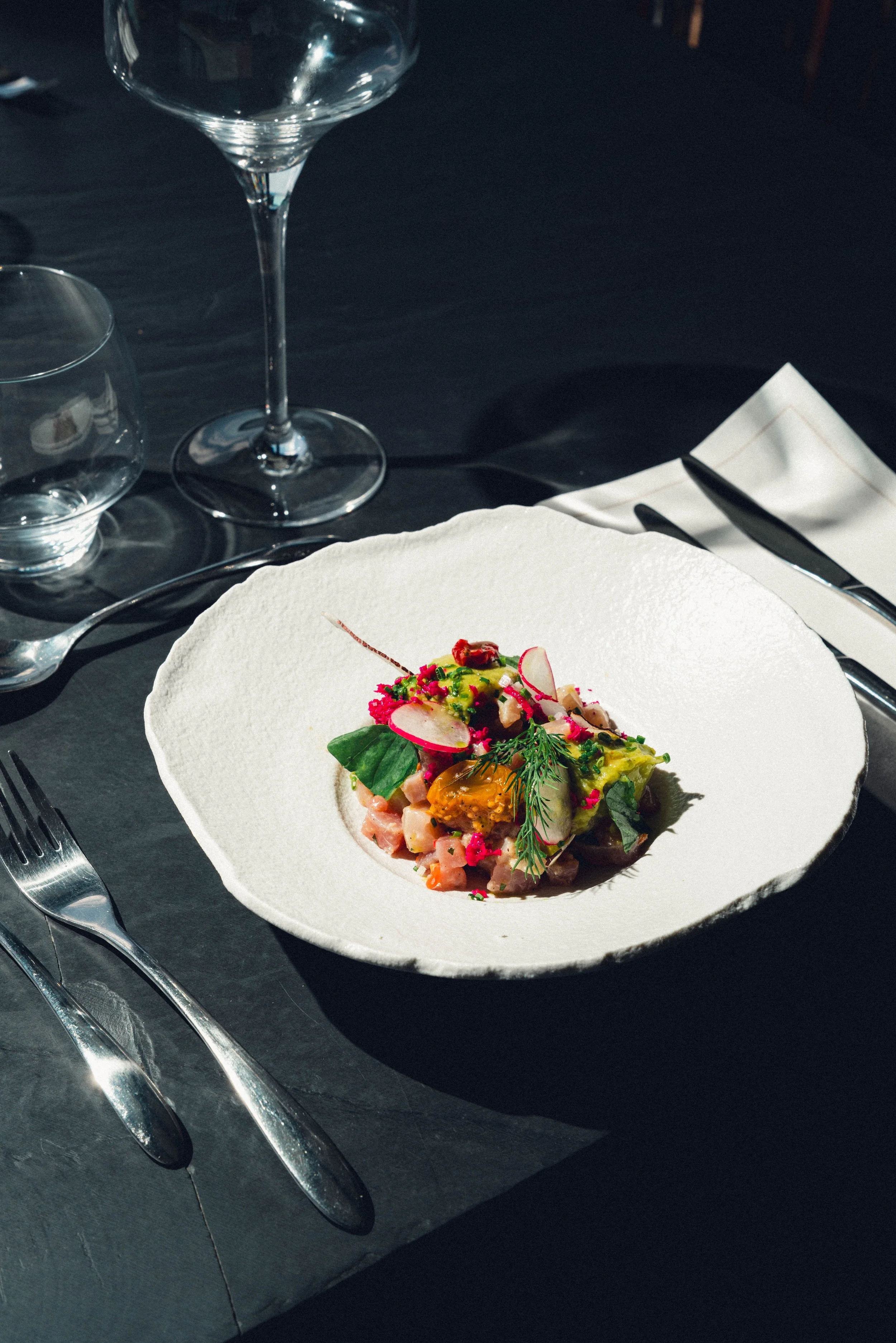 Assiette de tartare de poisson ornée de légumes, avec un verre à vin rouge, un verre d'eau et des couverts sur une table sombre, dans un restaurant élégant.