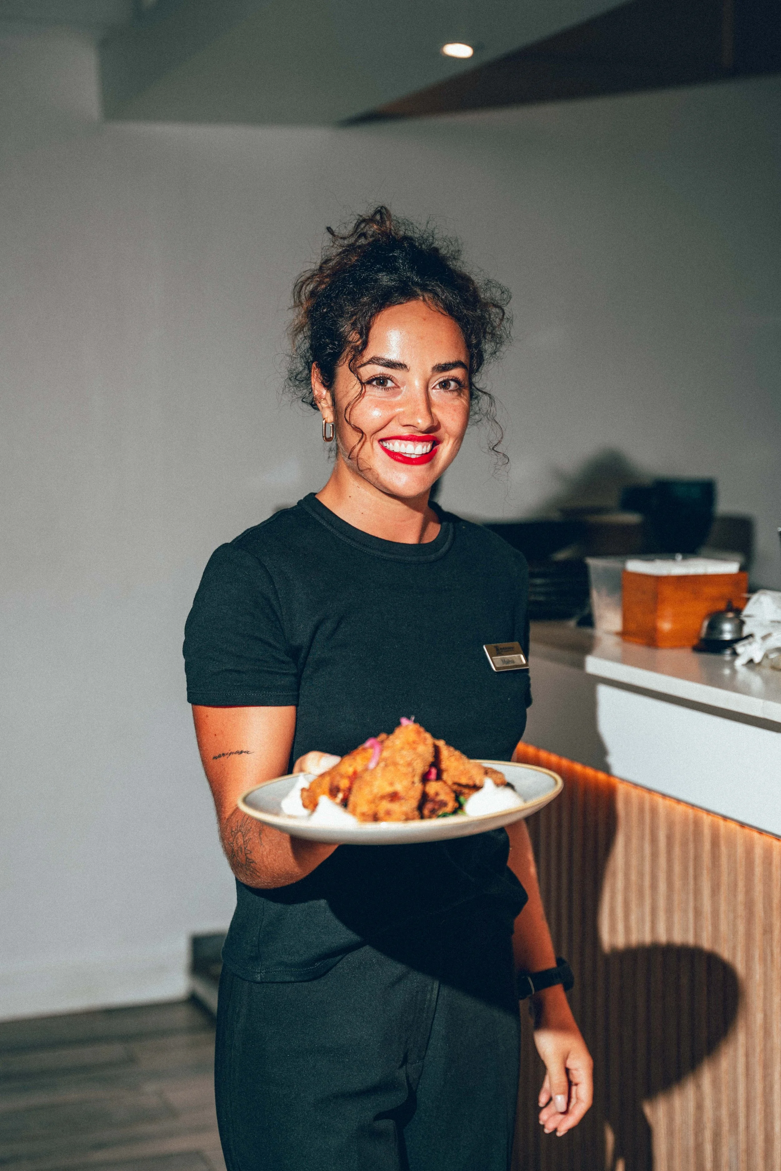 Photographie culinaire d'une employée de restaurant souriante avec des cheveux bouclés, portant un uniforme noir, présente un plat de nourriture. Elle porte un badge et se trouve dans un intérieur moderne.