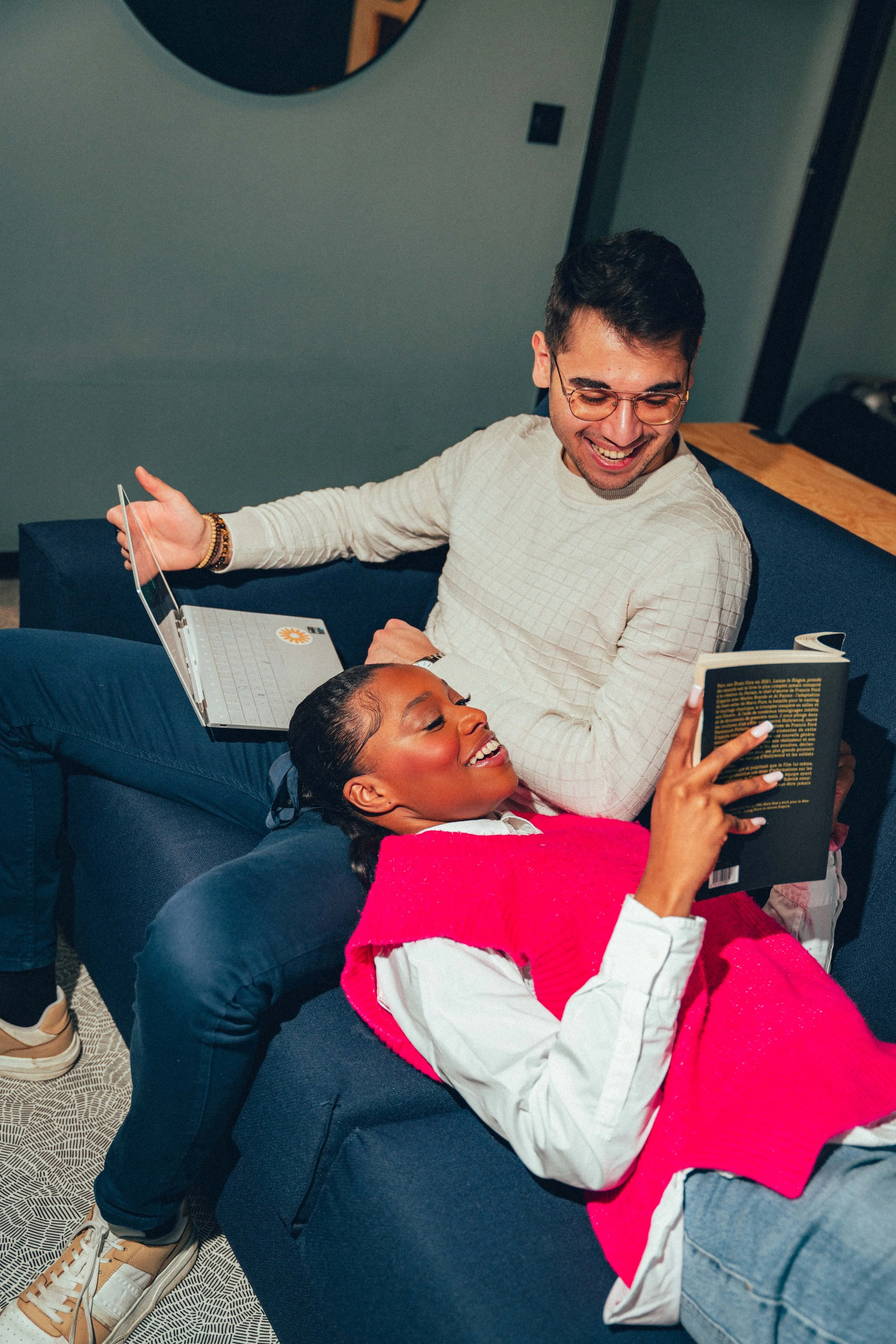 Deux personnes, un homme avec des lunettes et une femme, se détendent et rient ensemble sur un canapé, lisant un livre. La scène se déroule dans un intérieur moderne.