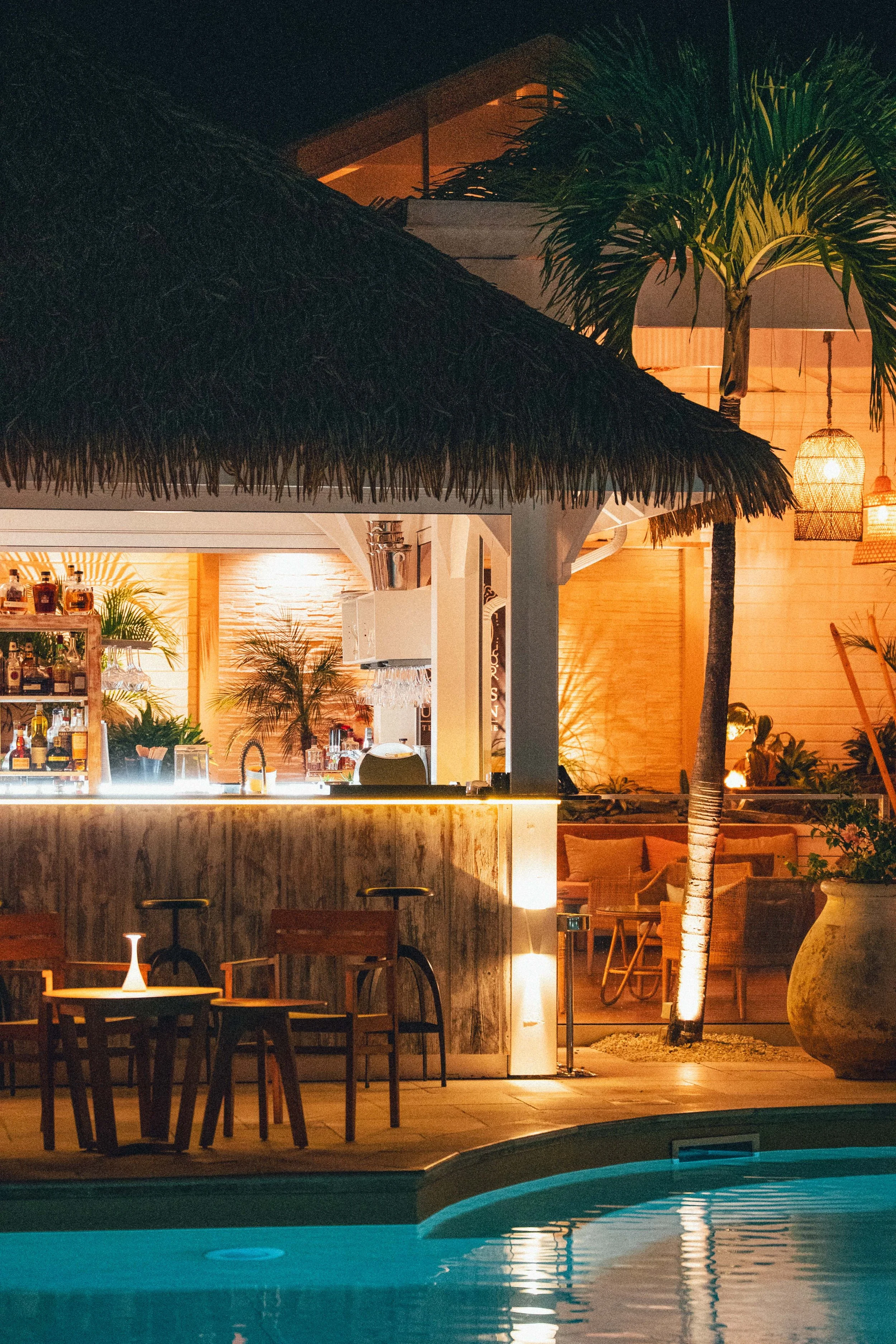 Piscine éclairée de nuit avec un bar en bois, des chaises en bois, une paillote en toit de chaume, des lampes suspendues, un palmier et mobilier exotique dans un espace extérieur tropical