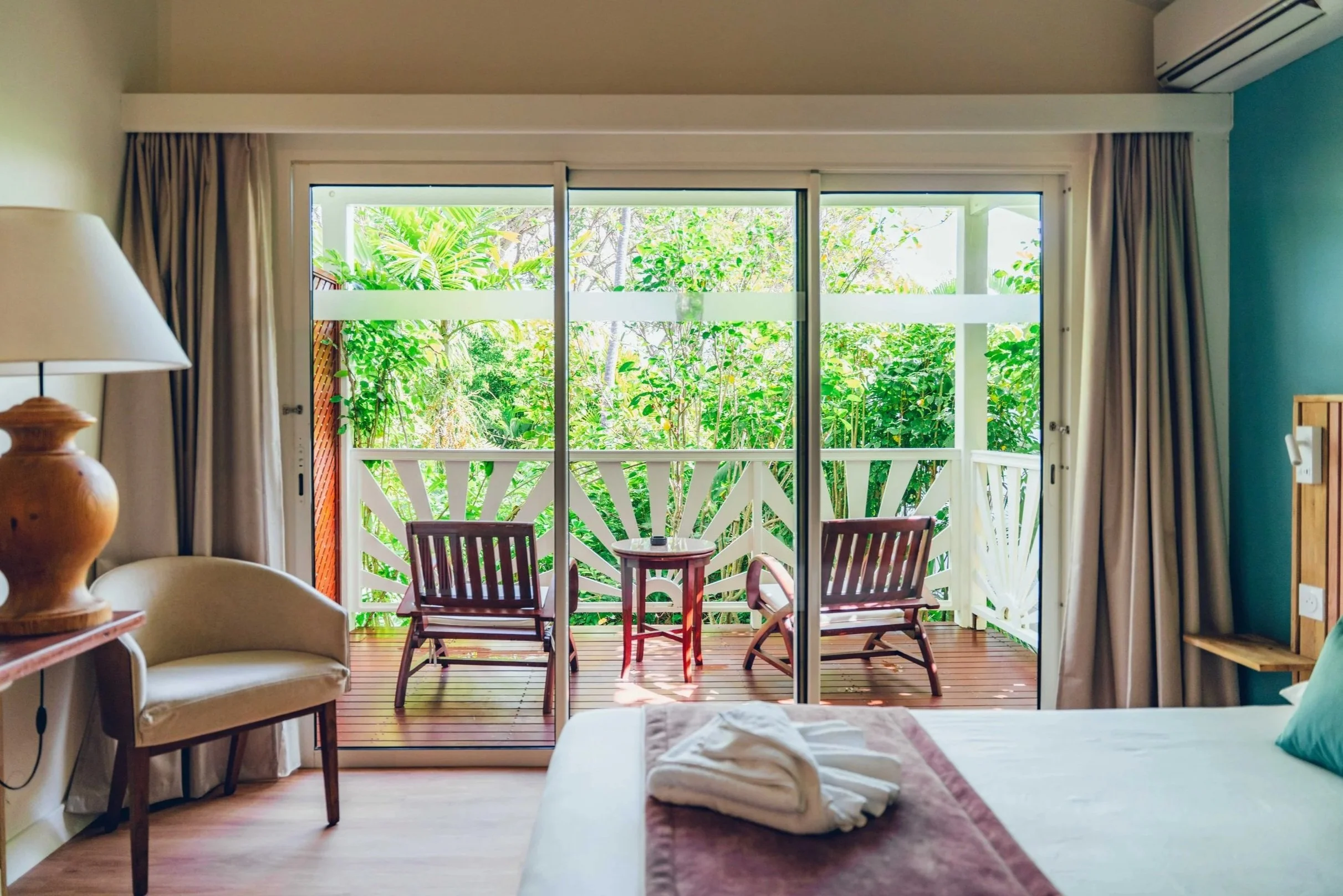 Vue intérieure d'une chambre d'hôtel avec un lit à gauche, une chaise en bois, un petit balcon avec deux chaises en bois et une table, et une vue verdoyante à travers des grandes fenêtres.