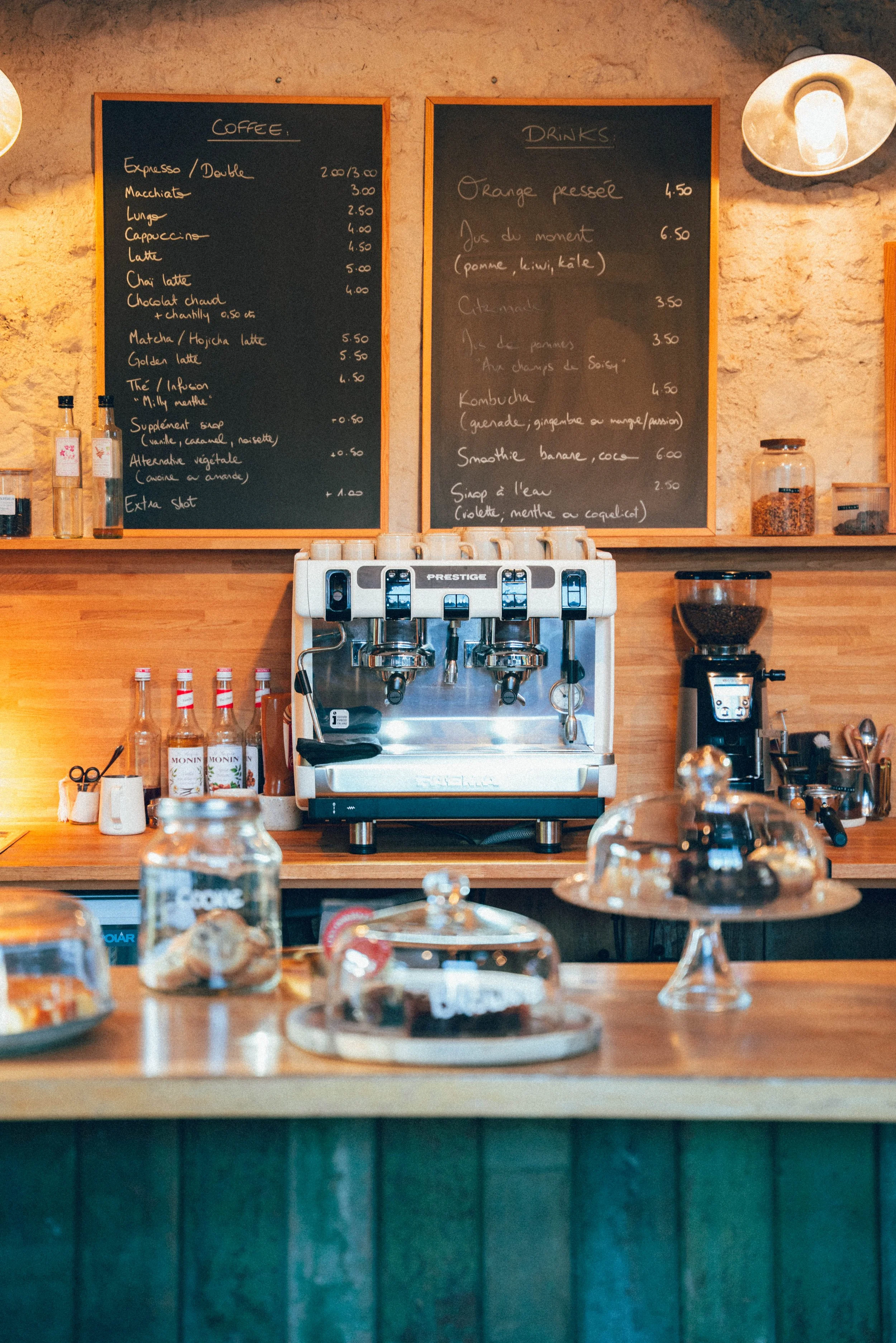 Un comptoir de café avec une machine à espresso, des bouteilles de sirop, et des gâteaux sous des cloches en verre. Deux grands tableaux noirs avec le menu écrit en français sont au mur en arrière-plan.