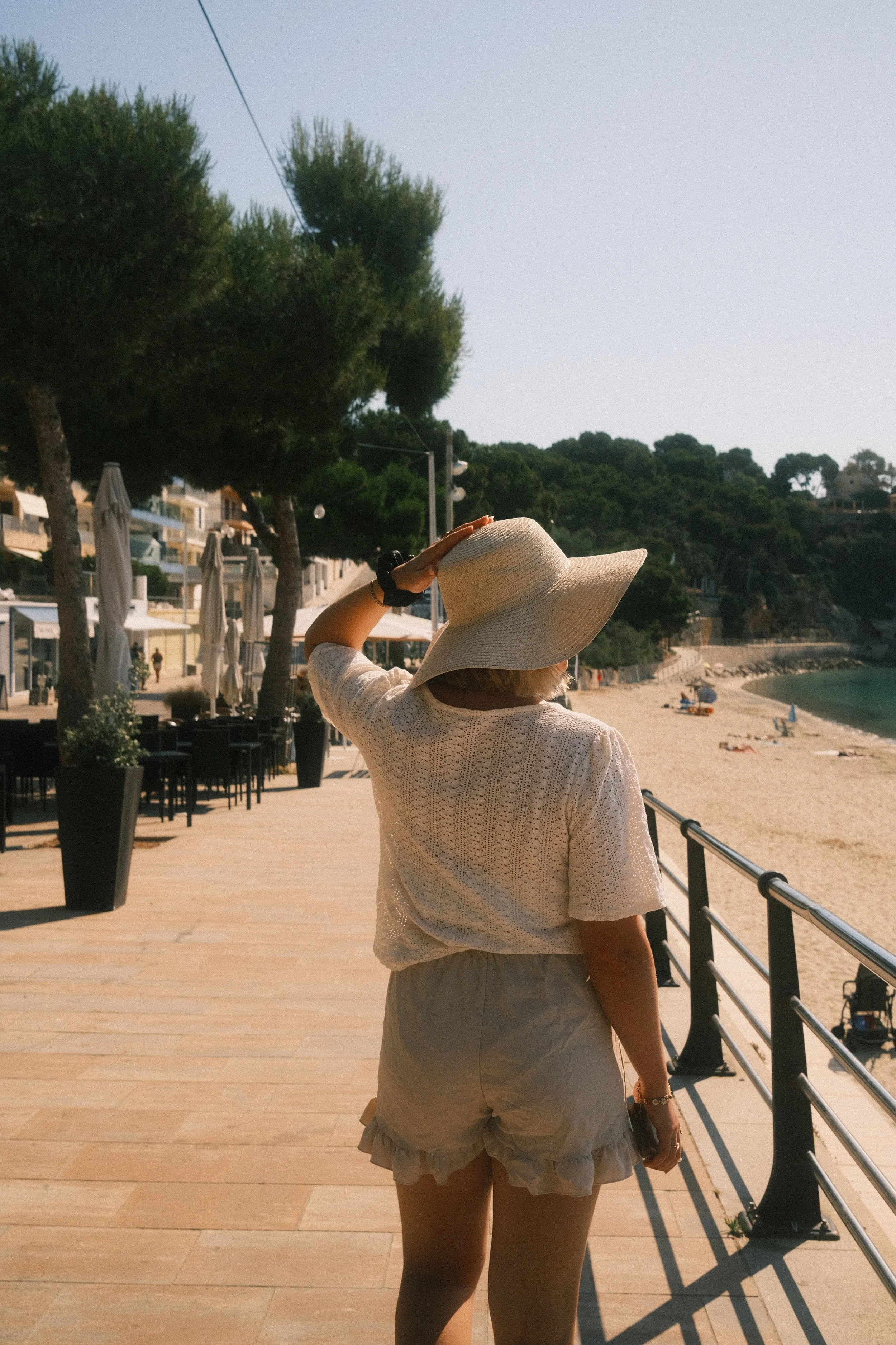 Une femme portant un chapeau de soleil blanc marche le long d'une promenade en bord de mer, avec des parasols et des bâtiments en arrière-plan.
