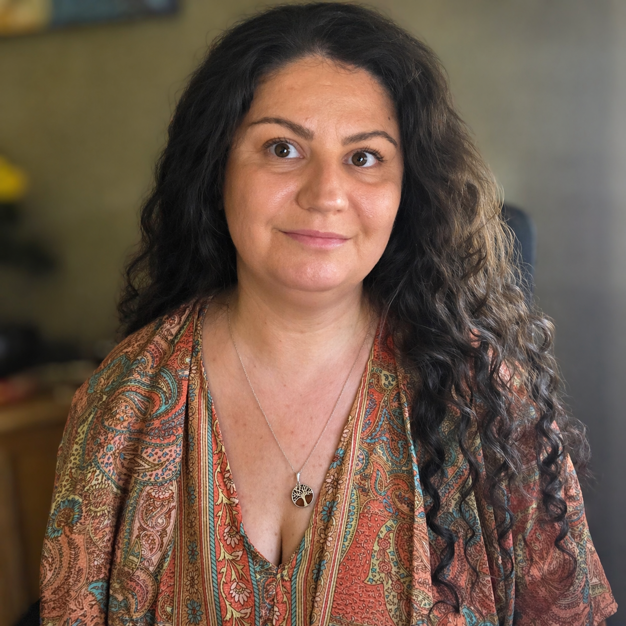 A woman with long curly dark hair, wearing a patterned top and a silver tree of life necklace, looking at the camera with a neutral expression.