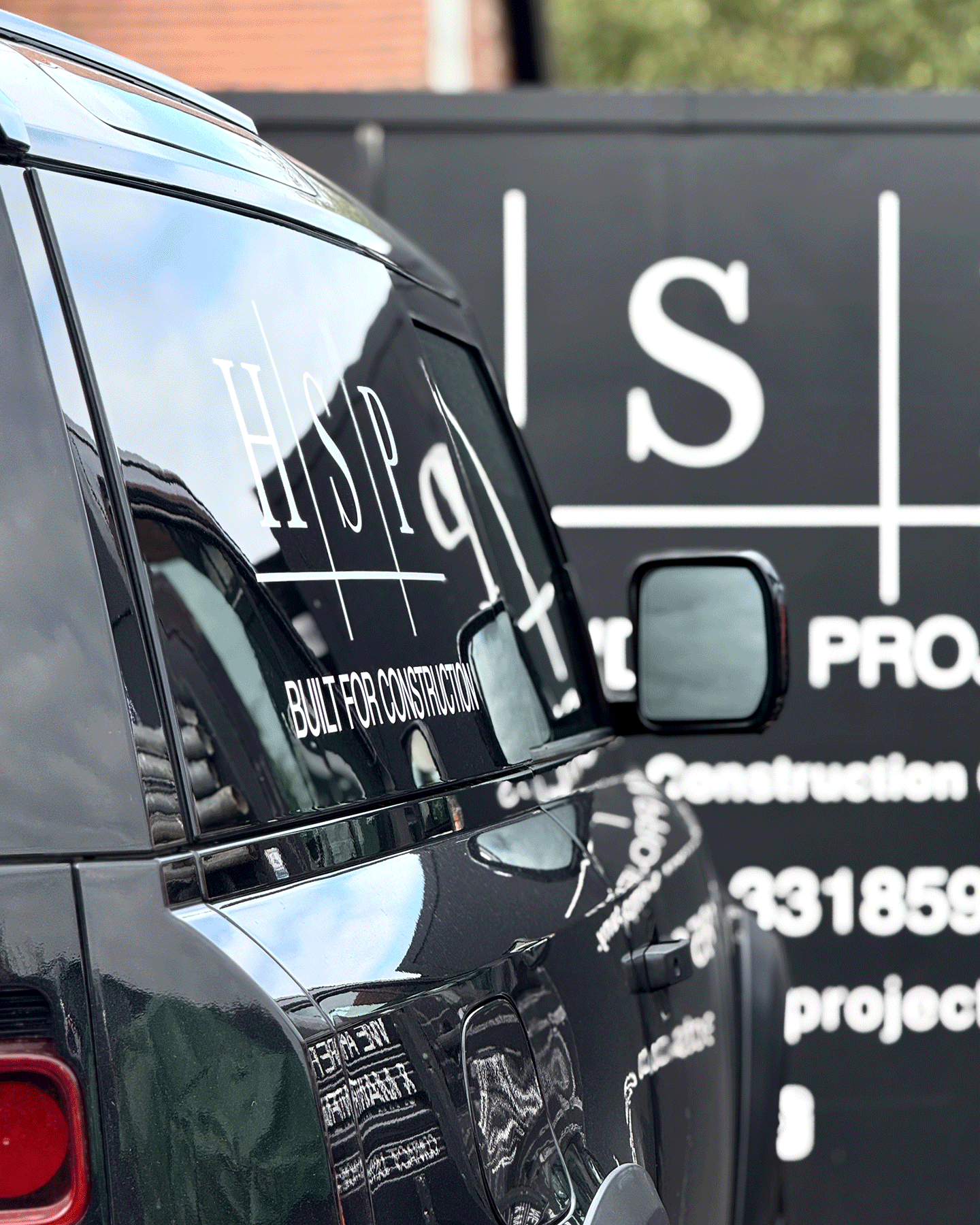 Close-up of the back of a black SUV with white lettering on the side, parked beside a black sign with white text and a logo, with a brick building and some trees in the background.
