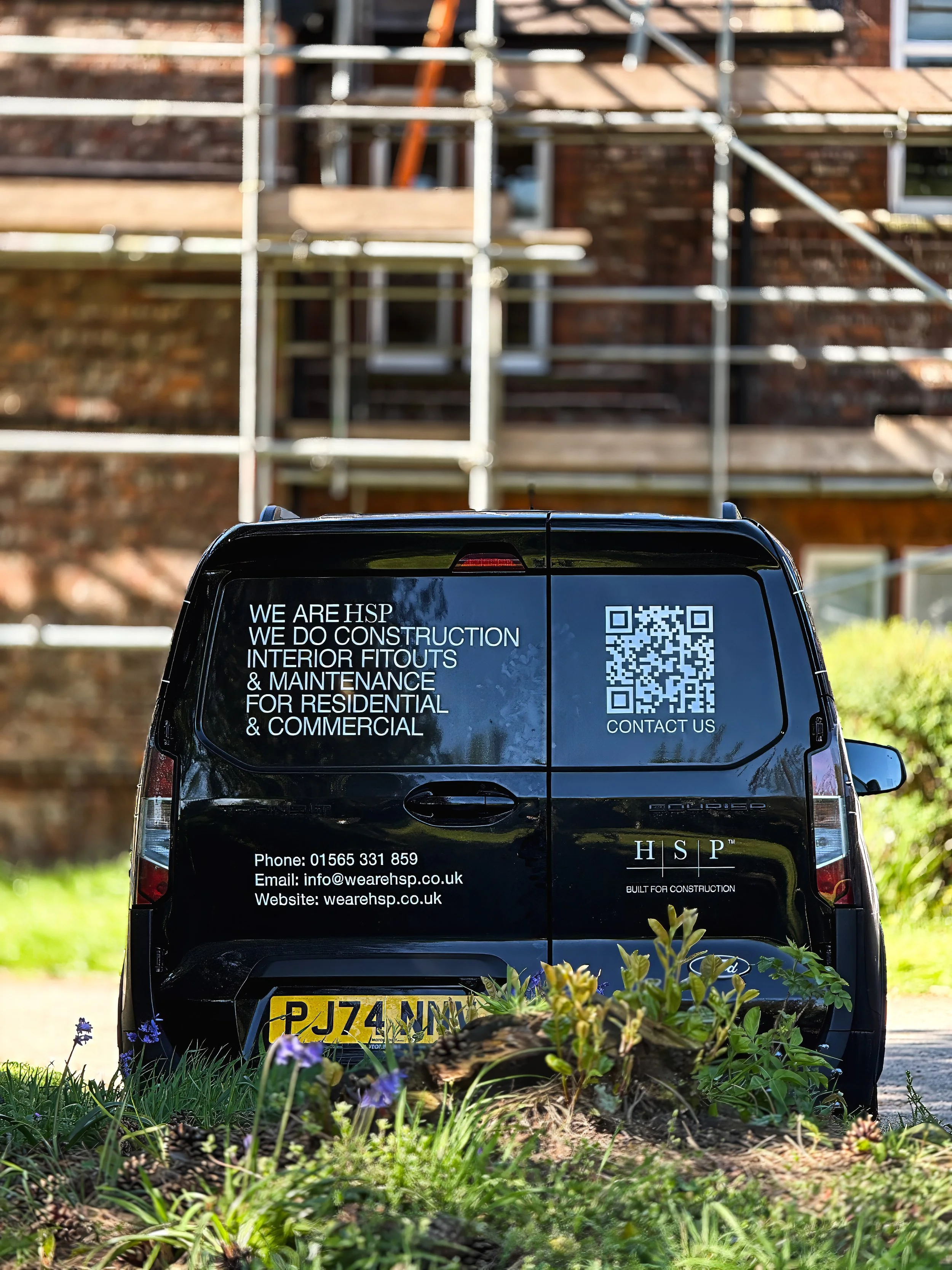 Back of a black van with construction company details, parked in front of a house with scaffolding. The text mentions construction, interior fit-outs, maintenance, and contact information.