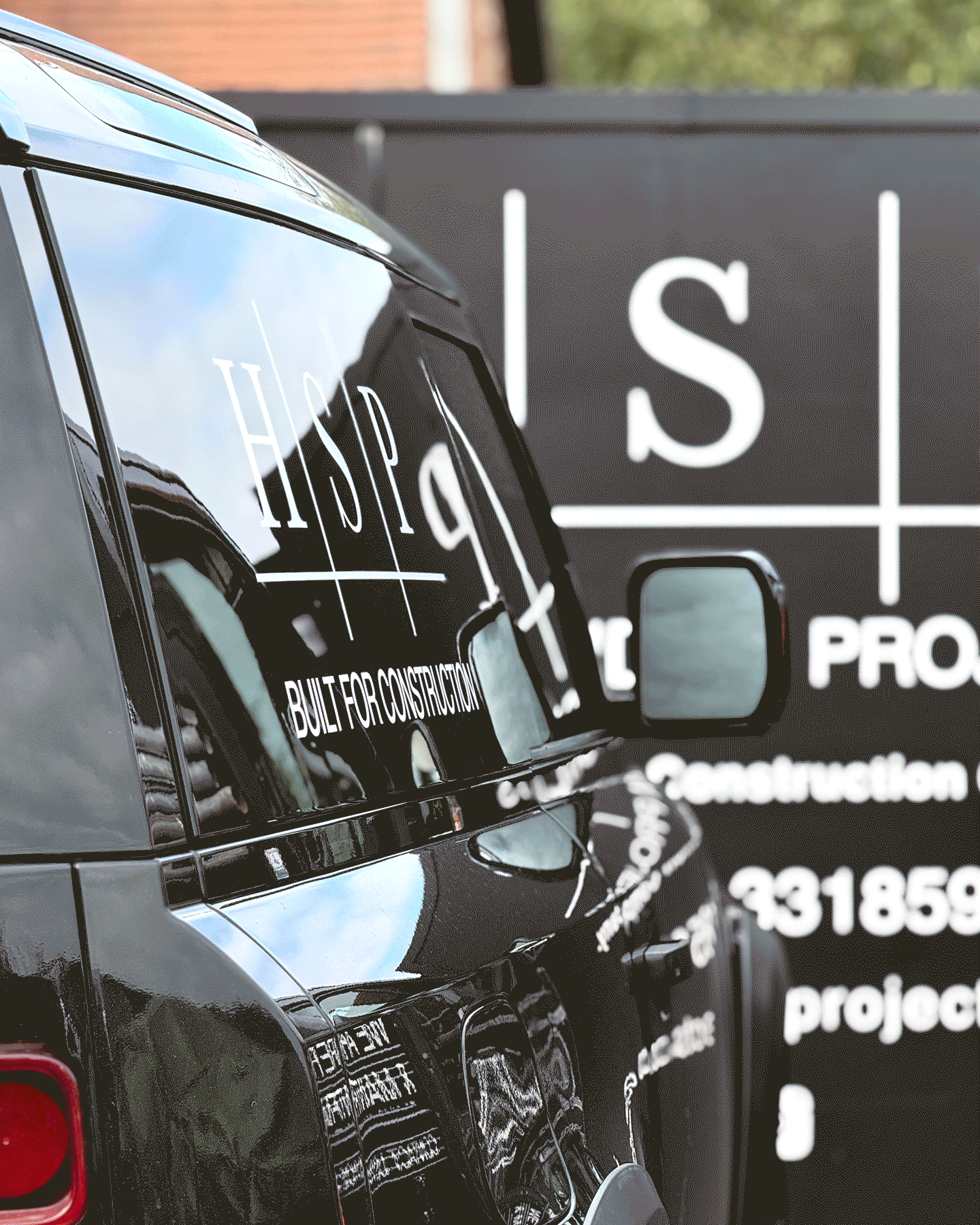 Close-up of a black construction company's vehicle with white lettering on the side, reflecting a nearby building, and a large black sign with white text in the background.