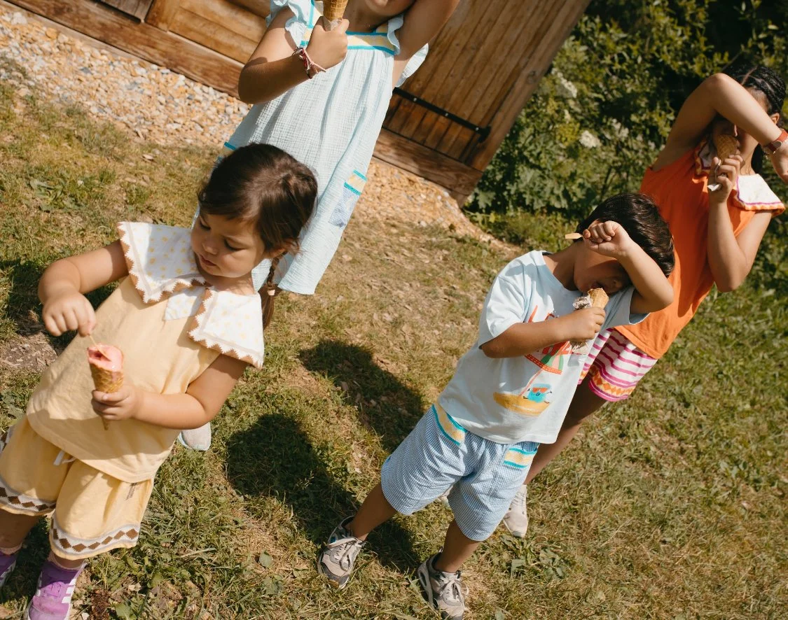 Children eating ice cream outdoors on a sunny day, standing on grass near a wooden fence.