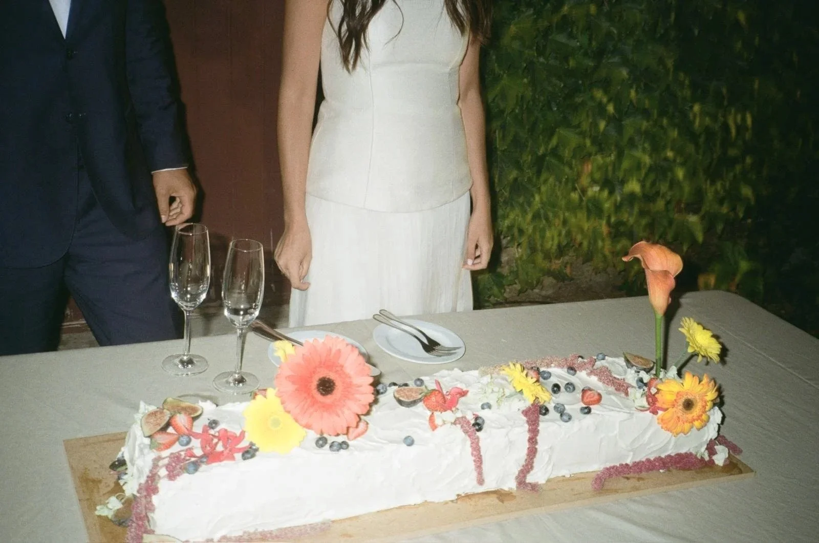 Bolo de casamento retangular comprido. Decorado com flores e frutas. Dois noivos atrás do bolo, a noiva com vestido branco e o noivo de fato, num ambiente ao ar livre.