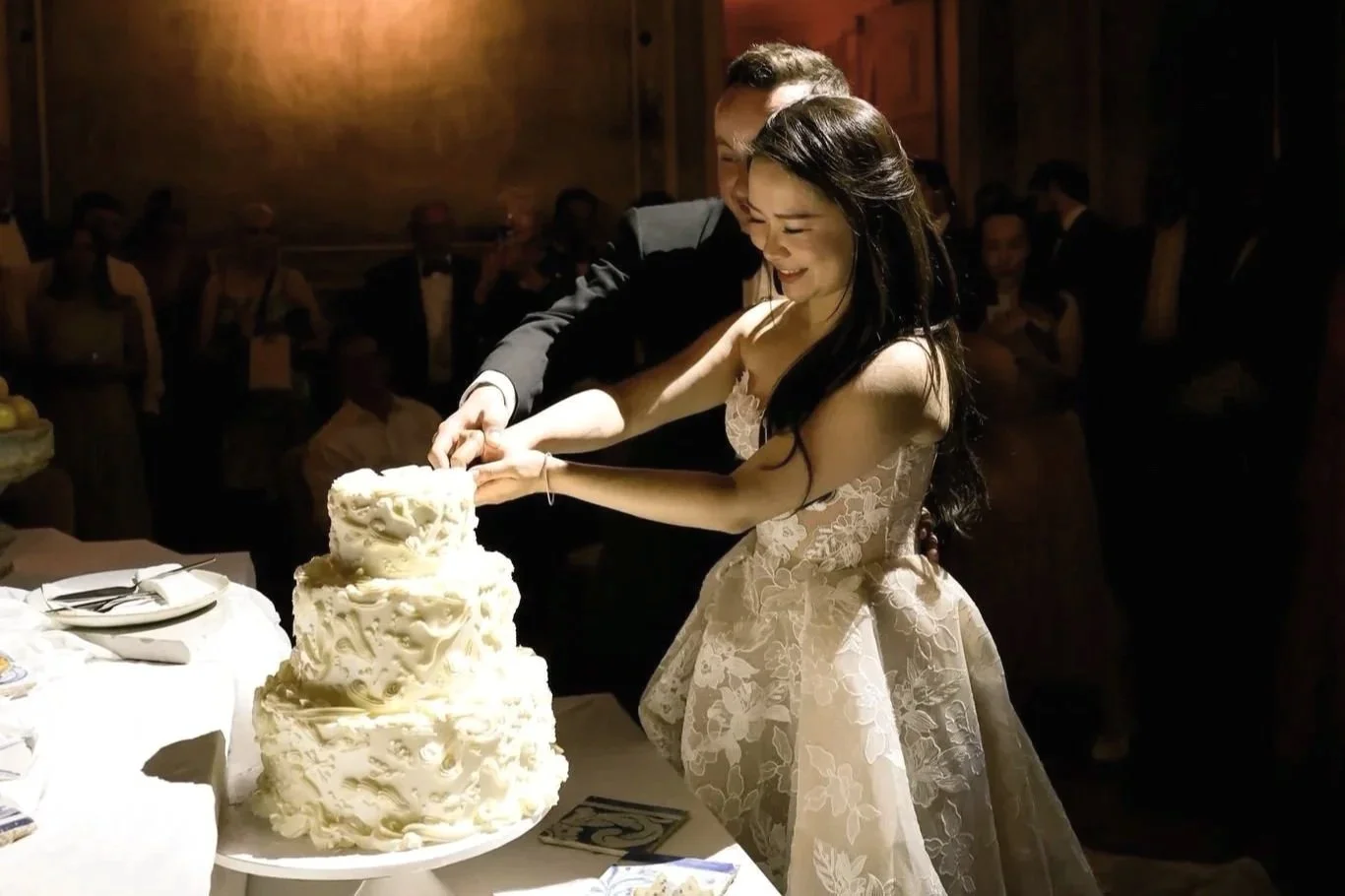 Casal de noivos cortando o bolo de casamento numa festa, com convidados ao fundo.