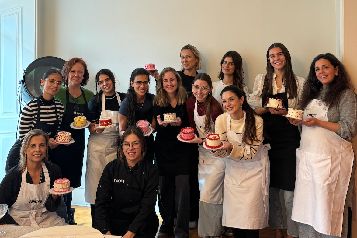 Grupo de mulheres sorrindo segurando bolos decorados em um ambiente bem iluminado, possivelmente em um curso de confeitaria.