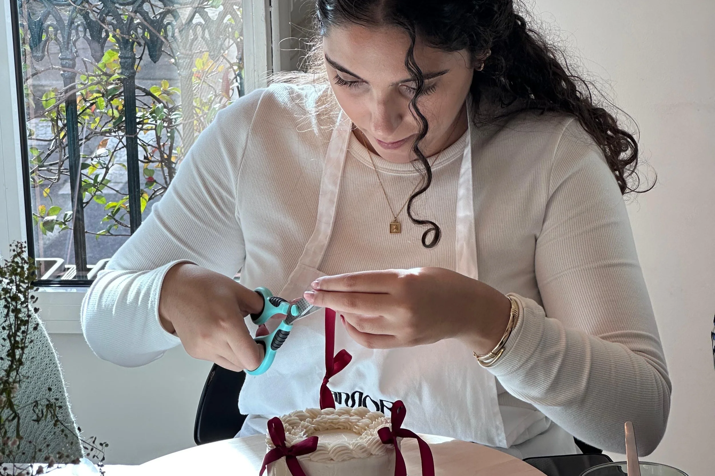 Rapariga a decorar um bolo durante um workshop promovido pela AMORI CAKES. A cortar fita vermelha com uma tesoura e com um pequeno bolo com laços vermelhos à sua frente.