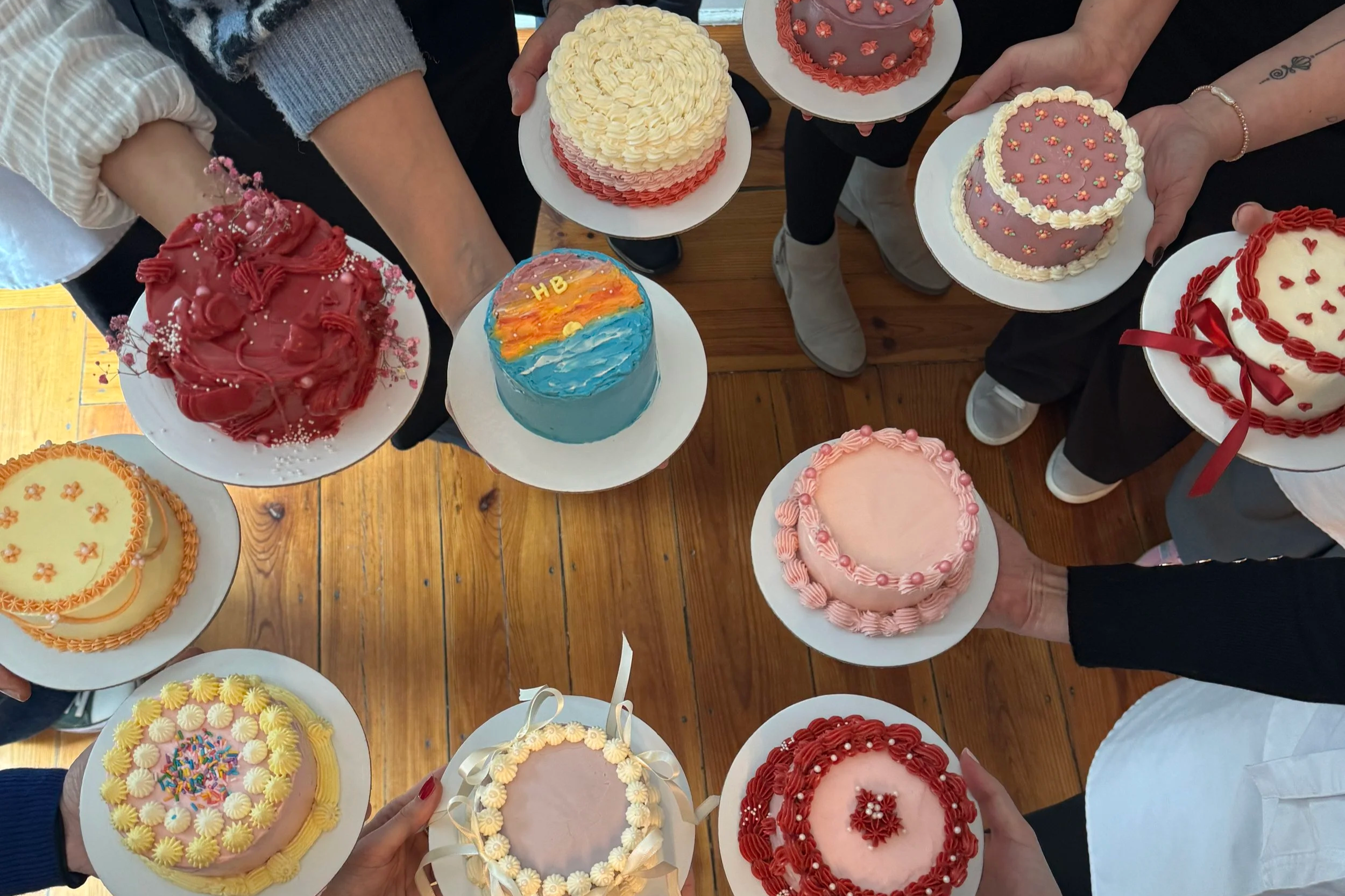 Vários bolos decorados com diferentes cores, temas e enfeites, segurados por várias pessoas, formando um círculo visto de cima. Os bolos têm detalhes decorativos variados, como flores, laços e outros desenhos.