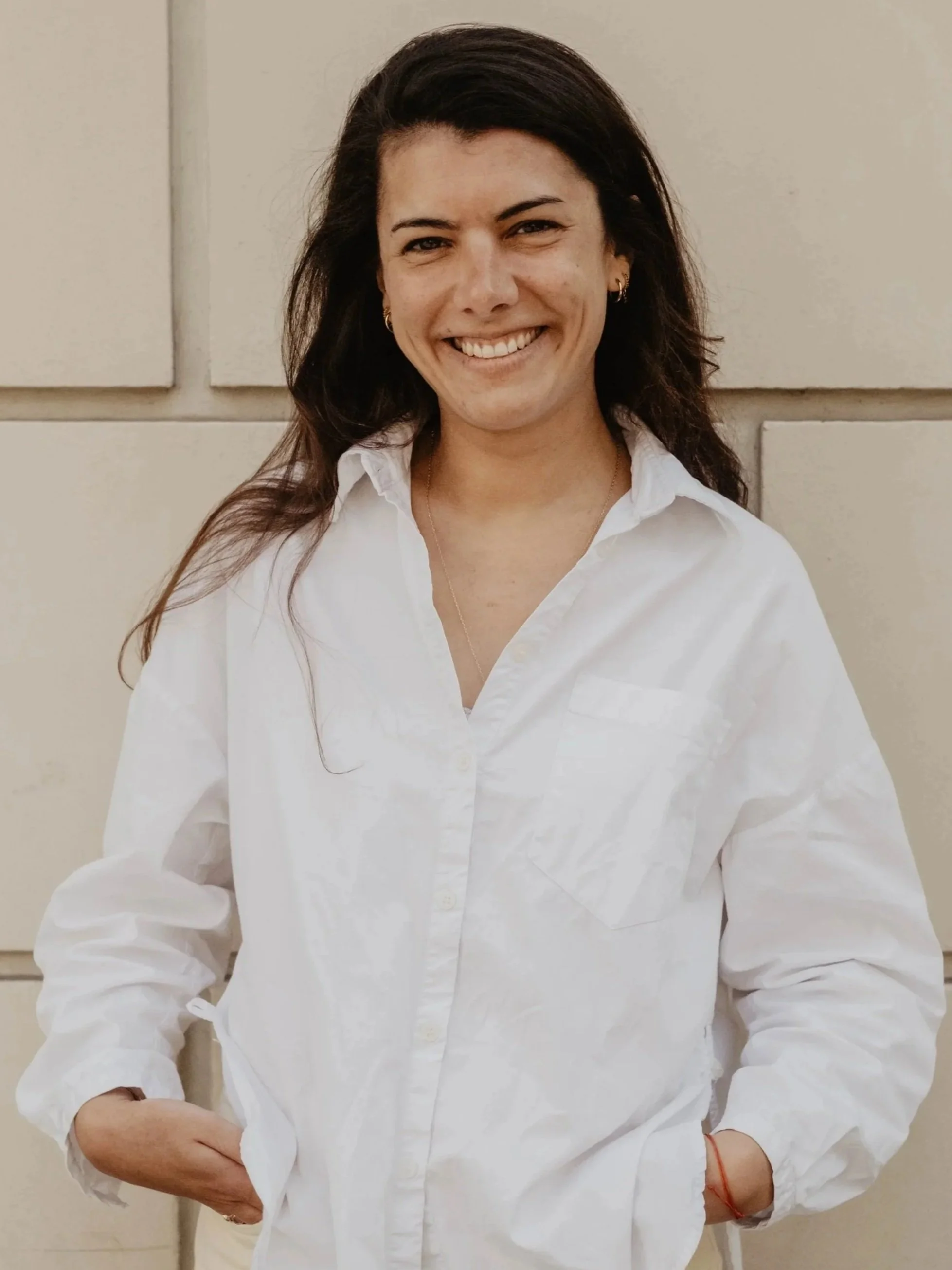 Une femme souriante portant une chemise blanche, les bras croisés, devant un mur beige.