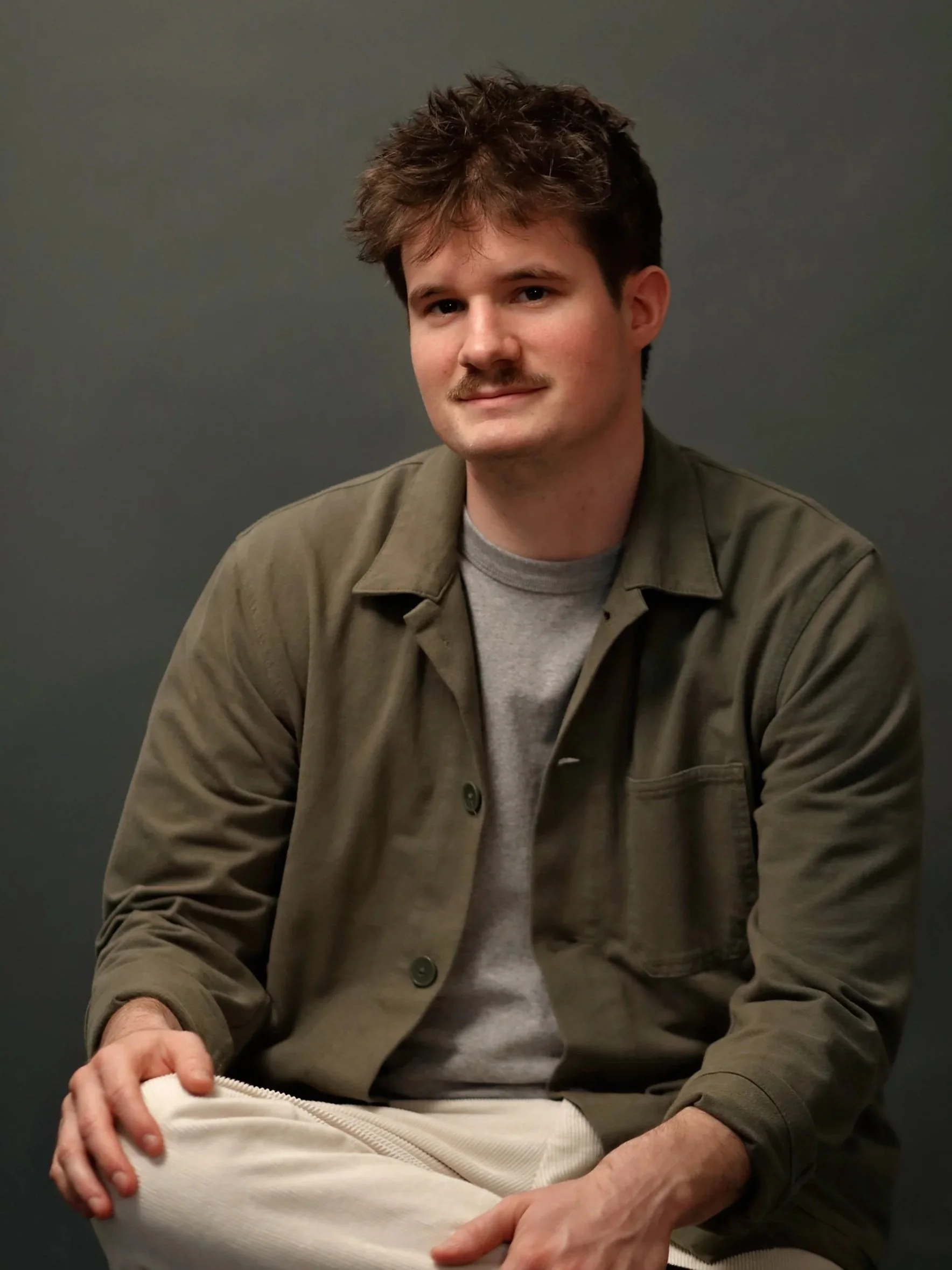 Un jeune homme avec des cheveux bouclés et une moustache, portant une chemise marron, assis contre un fond gris, souriant légèrement.