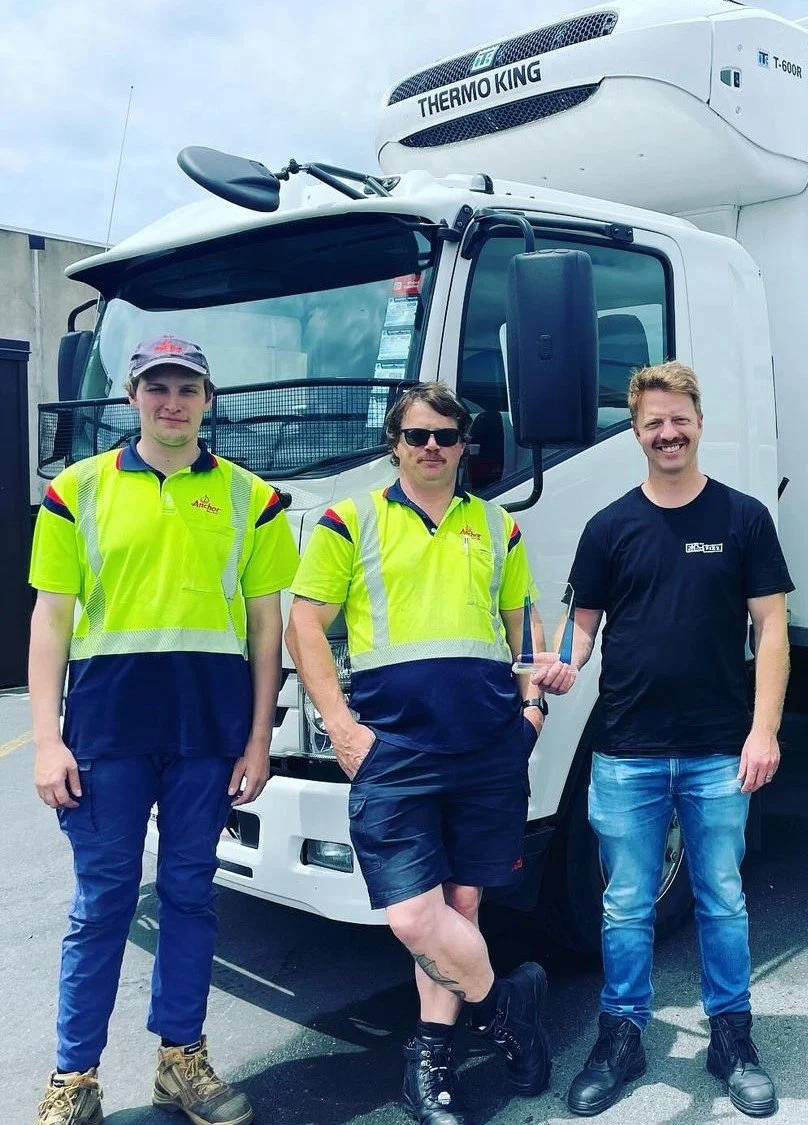 Three men standing in front of a truck, one holding an award. Two of the men are wearing high-visibility safety shirts, and one is in a black T-shirt. The truck has a Thermo King refrigeration unit on top.
