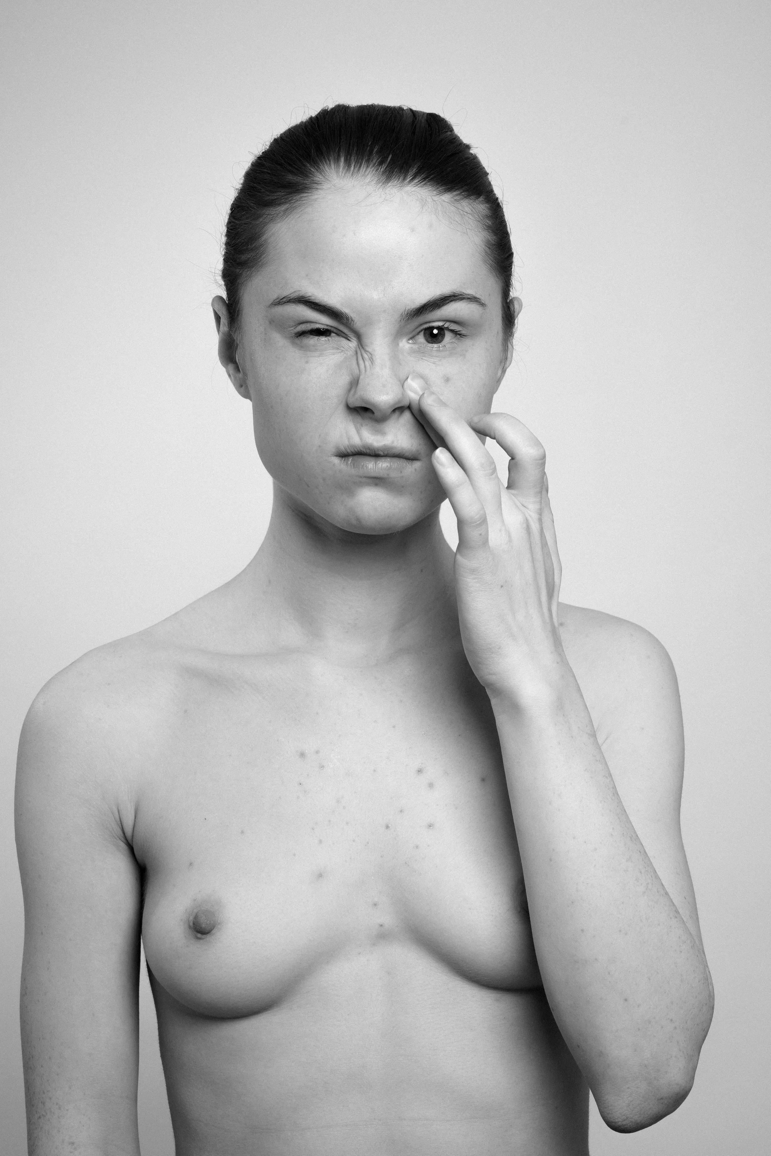 Studio portrait of a young woman in black and white. She manipulates her face, makes faces, grimaces, and touches her face with her left hand.