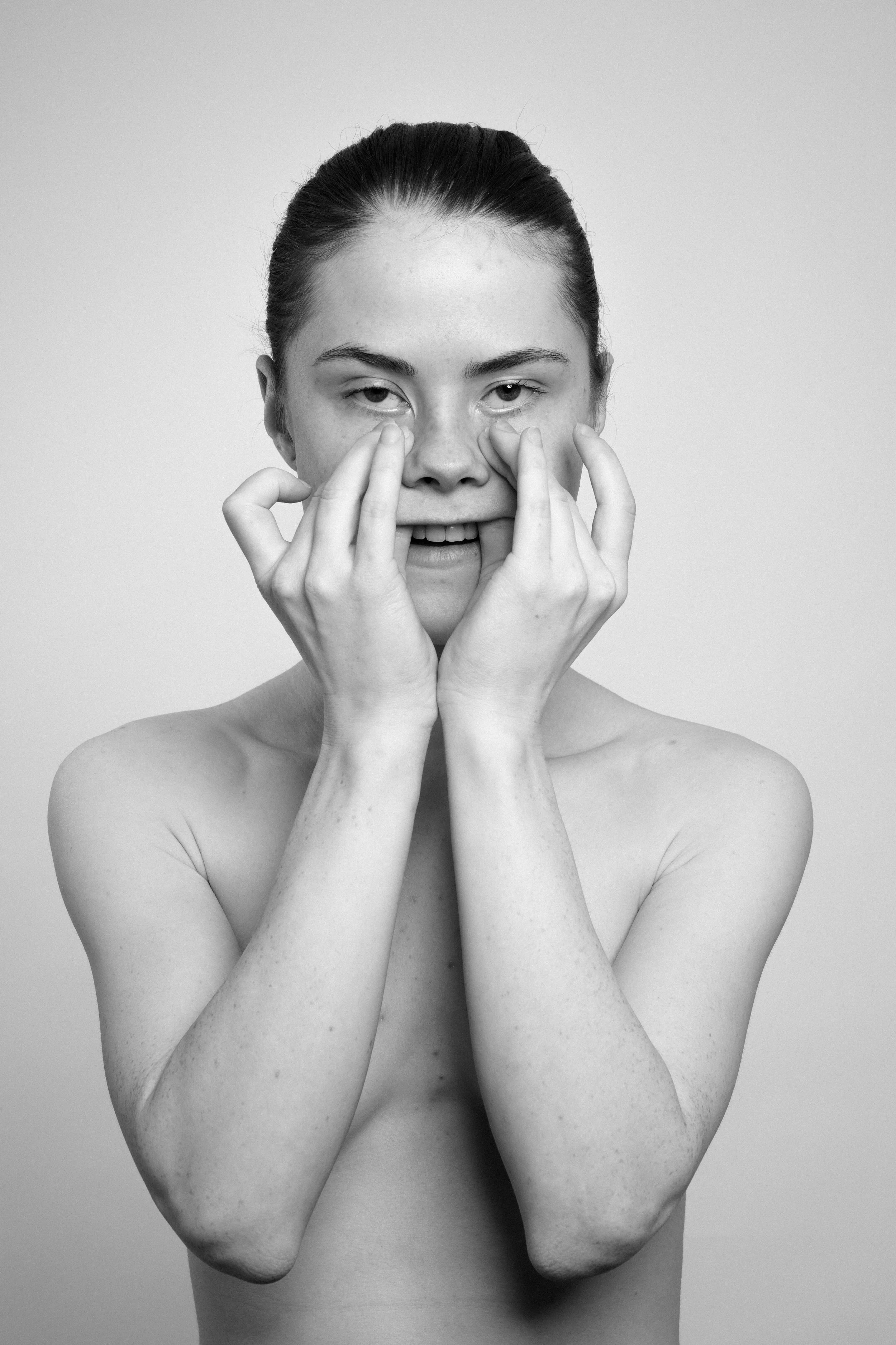 Studio portrait of a young woman in black and white. She manipulates her face, stretches her skin, with her both hands, makes faces, grimaces.