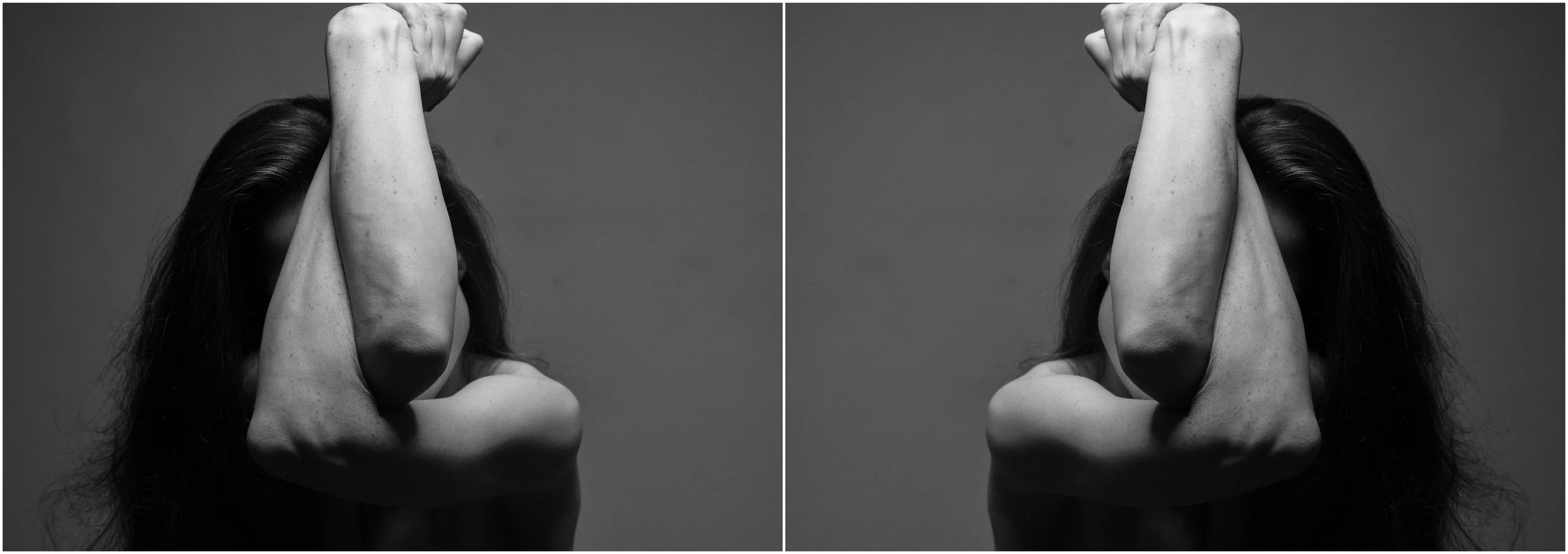A black and white mirror-reflection photographic portrait of a young woman. A close-up. She is covering her face with her arms crossed in front of her. Very strong shadows and contrasts.