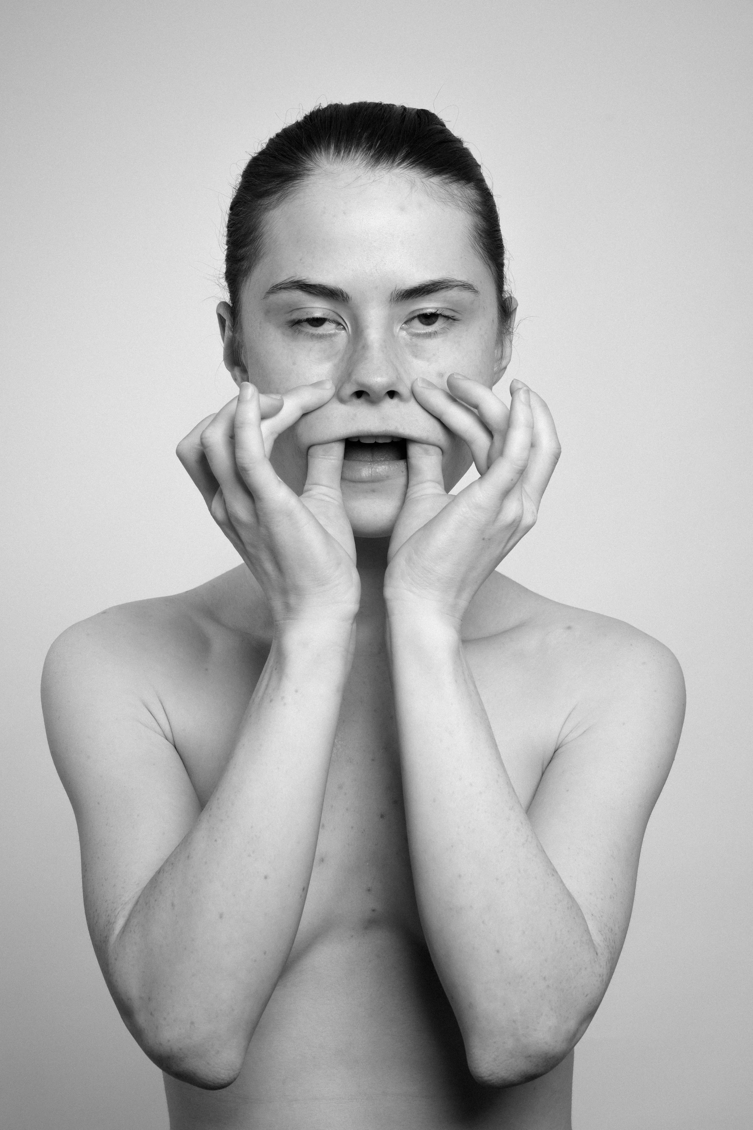 Studio portrait of a young woman in black and white. She manipulates her face, stretches her skin, with her both hands, makes faces, grimaces.