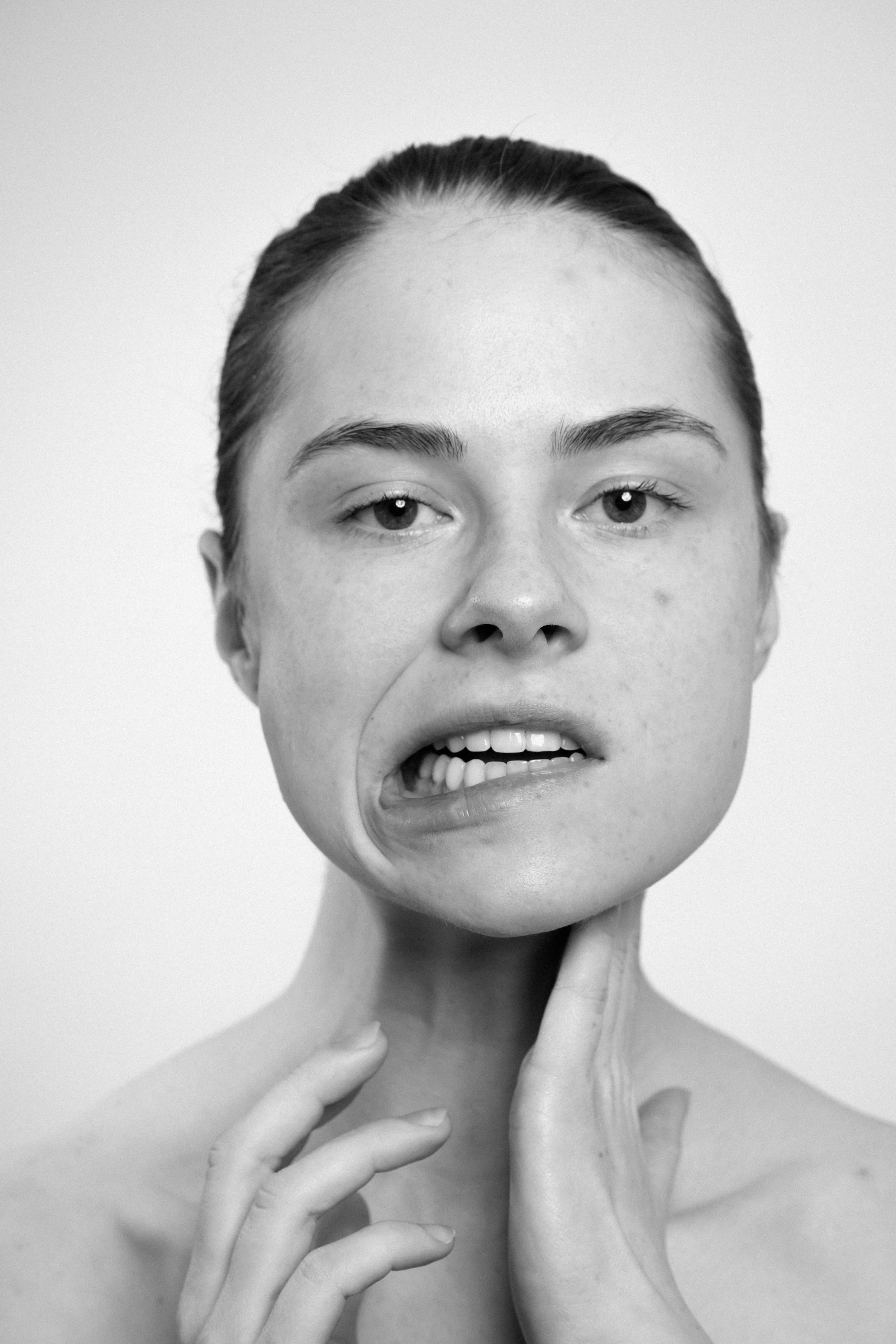 Studio portrait of a young woman in black and white. She touches her neck with her left hand. She makes faces, grimaces.