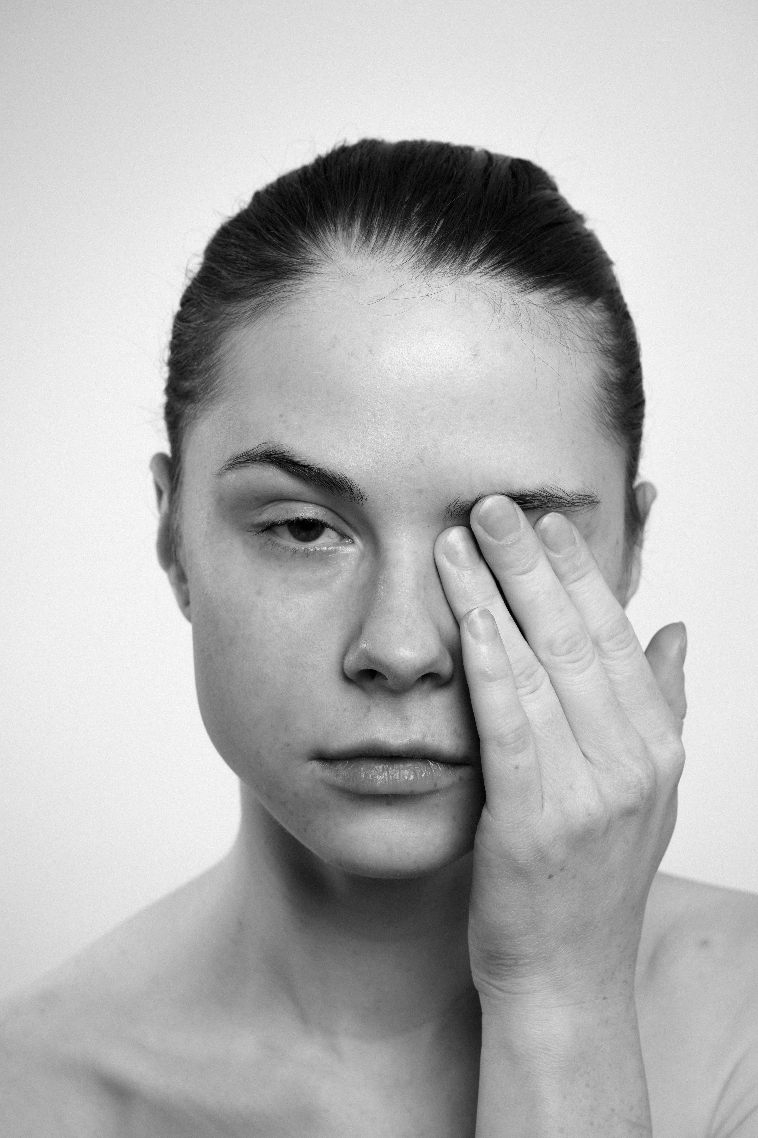 Studio portrait of a young woman in black and white. She covers up her eye with her left hand.