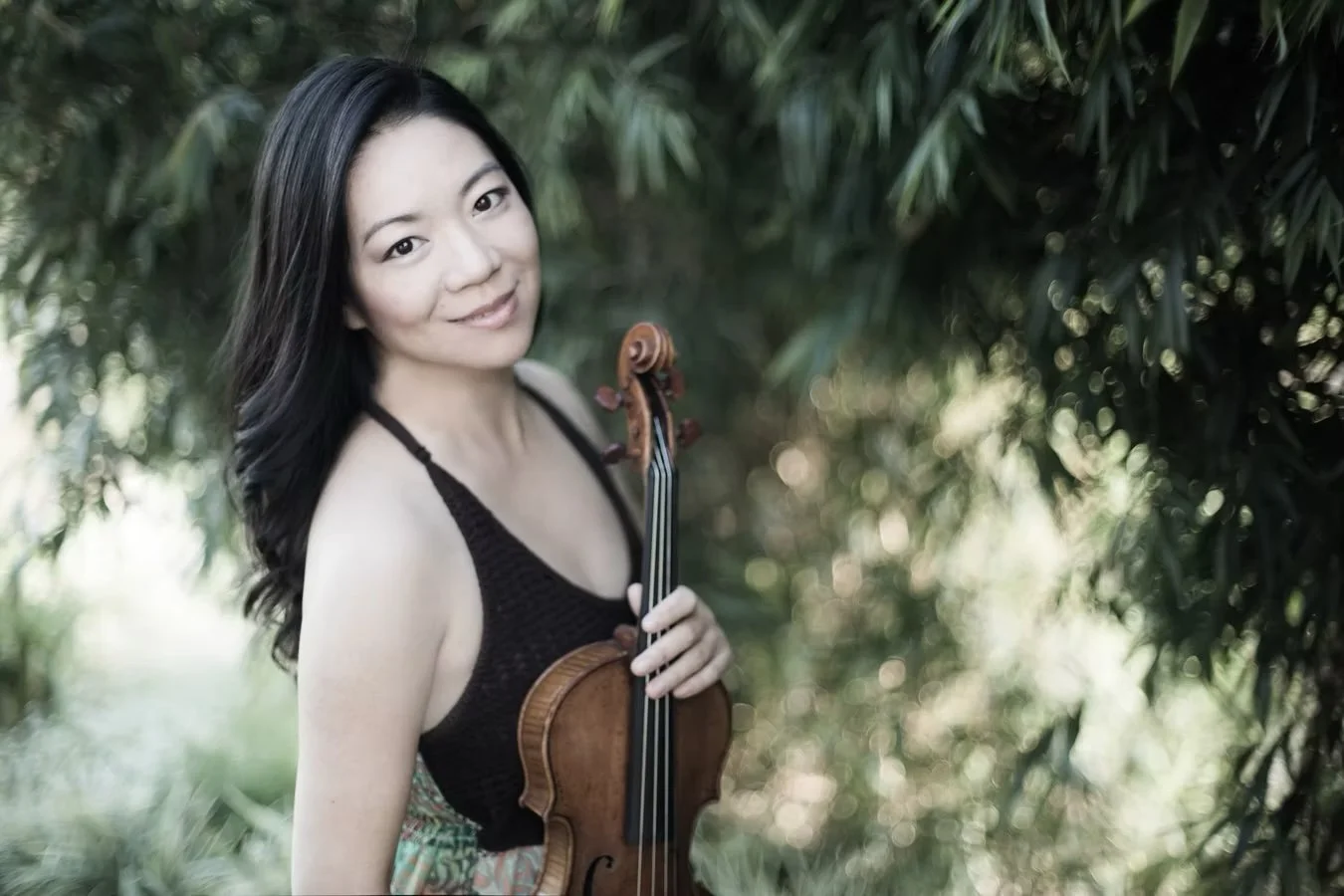 A woman with long dark hair smiling outdoors, holding a violin, with lush green foliage in the background.