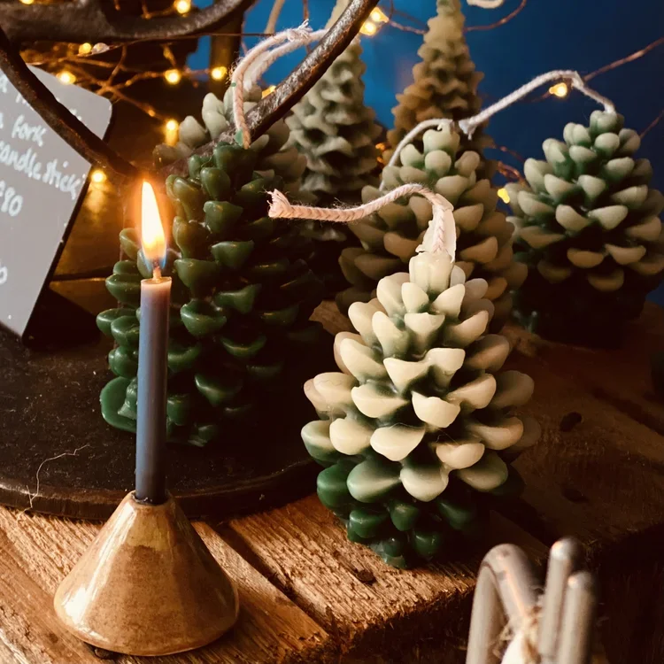Decorative display of small pinecone-shaped candles in green and white, arranged on a wooden surface with a black candle holder with a lit black candle in the foreground, and warm string lights in the background.