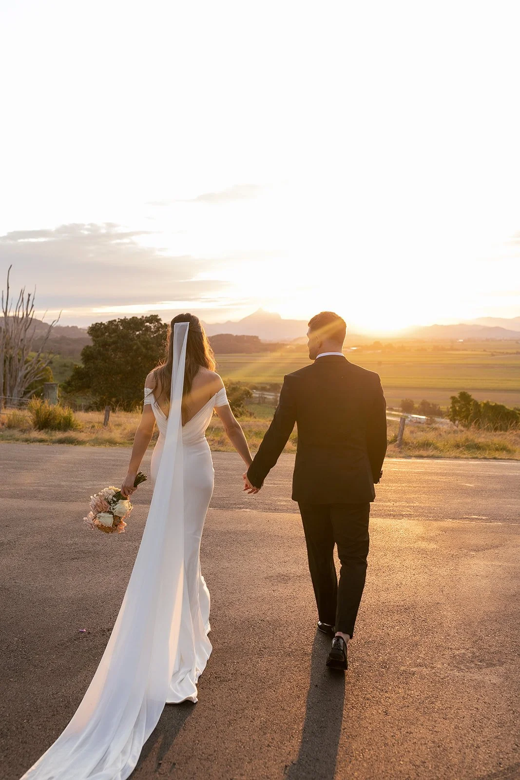 Bride and groom holding hands walking on an empty road during sunset, countryside landscape in the background.